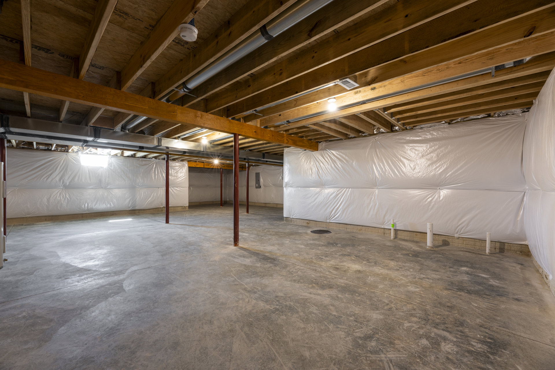White wall and ceiling with exposed wooden beams, metal pipes, light bulb fixture, and white plastic coverings along wall and ceiling.