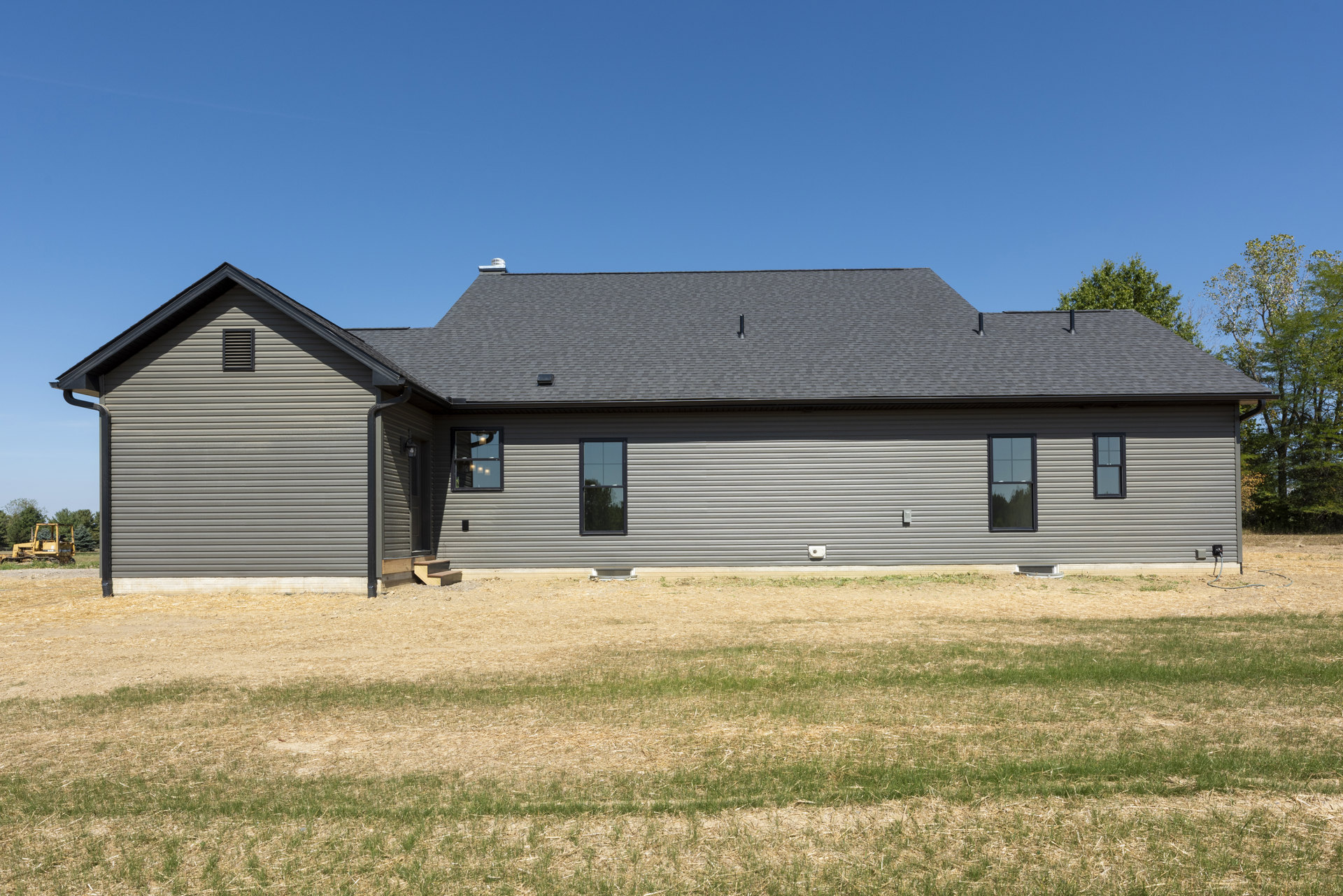 Grey farmhouse with black-framed windows, vented siding, grassy yard, metal barn in background, yellow bulldozer parked on lawn.