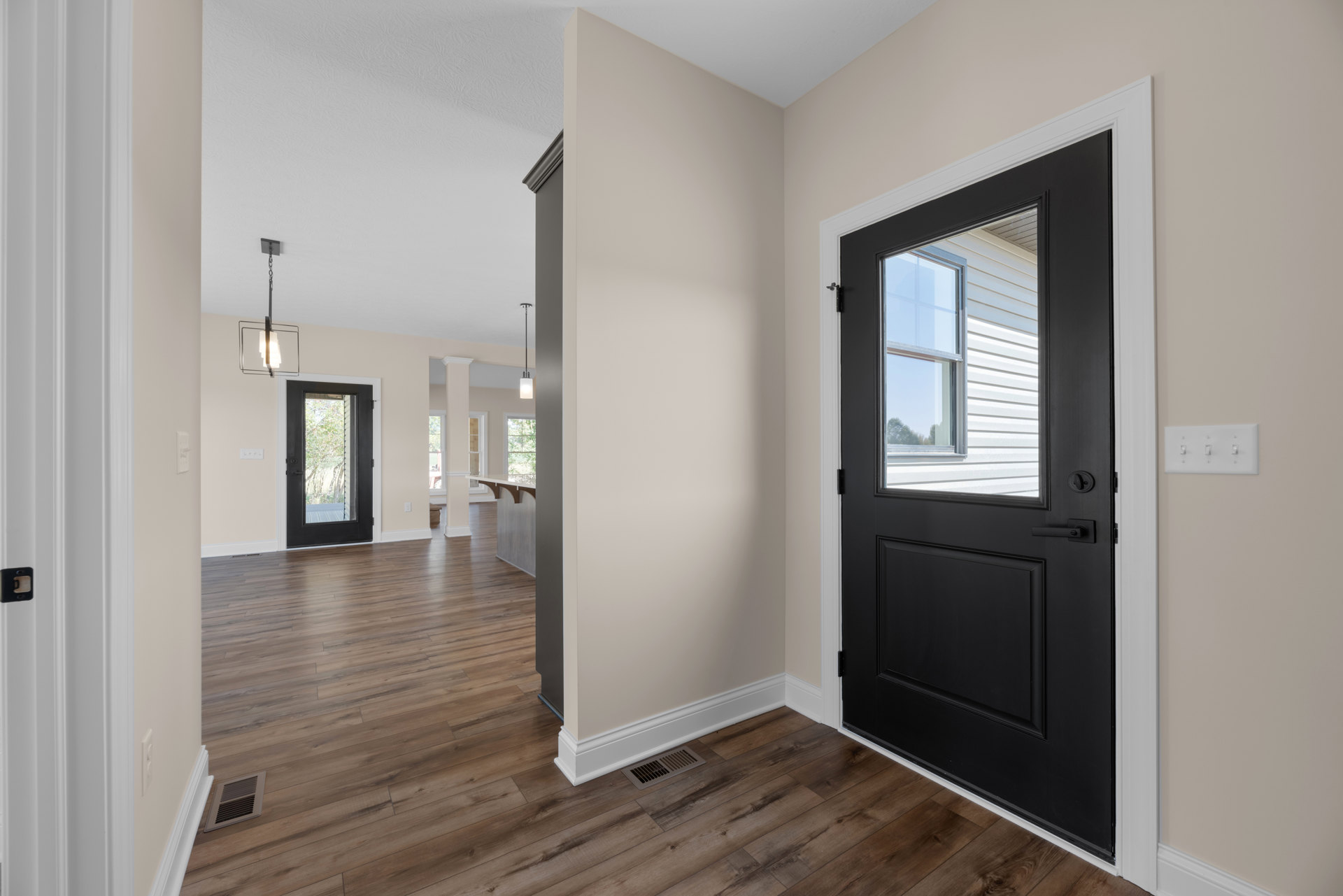 Black door with glass window, white walls, wood flooring, floor vent, row of light switches, window showing trees outside.