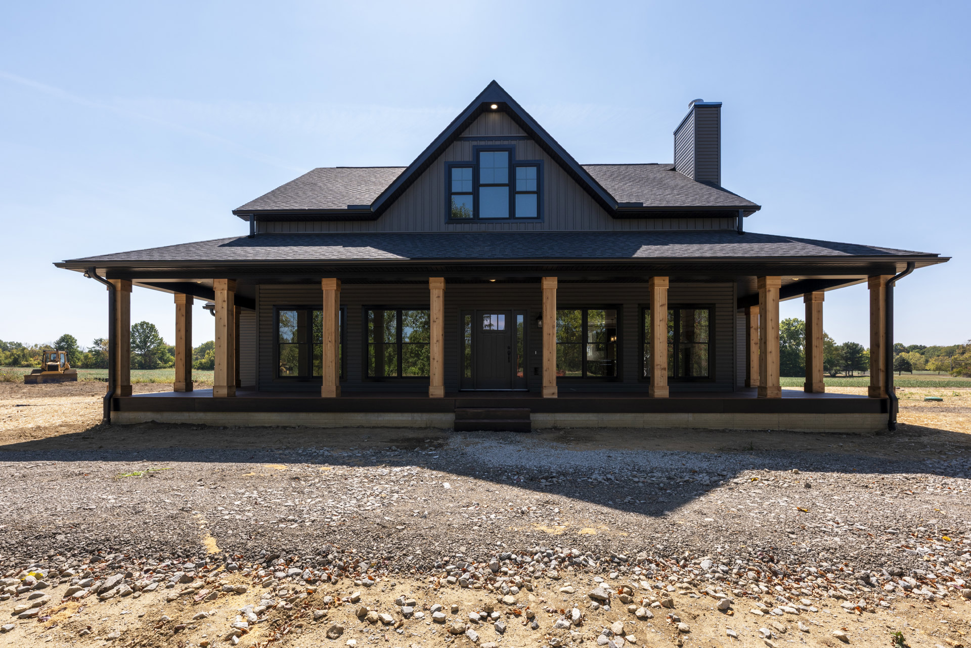 Wide front porch with white railings, dark wood door, multiple windows, stone landscaping, mature tree in background, blue sky overhead
