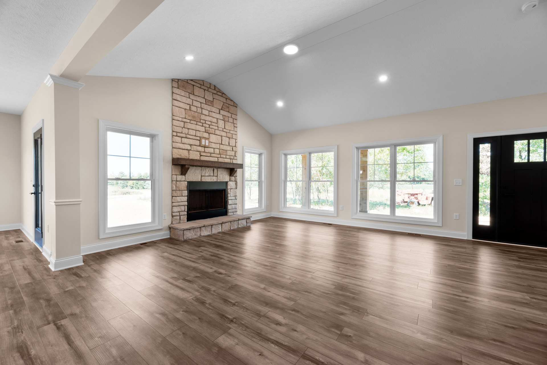 Spacious living area with hardwood flooring, stone fireplace with glass doors, large windows offering views of a field and tractor, and a door with glass panels overlooking trees.