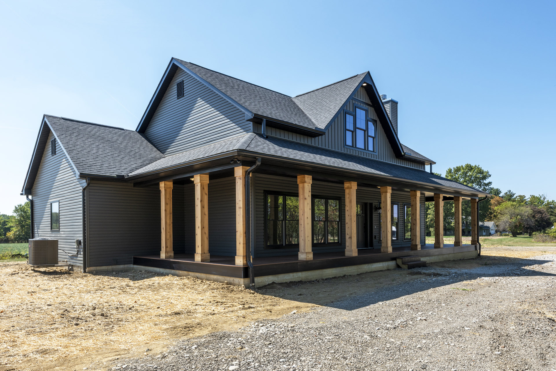 Wide front porch with white columns, wood decking, and a gabled roof; large windows framed by trim; mature trees and landscaped yard visible around the house