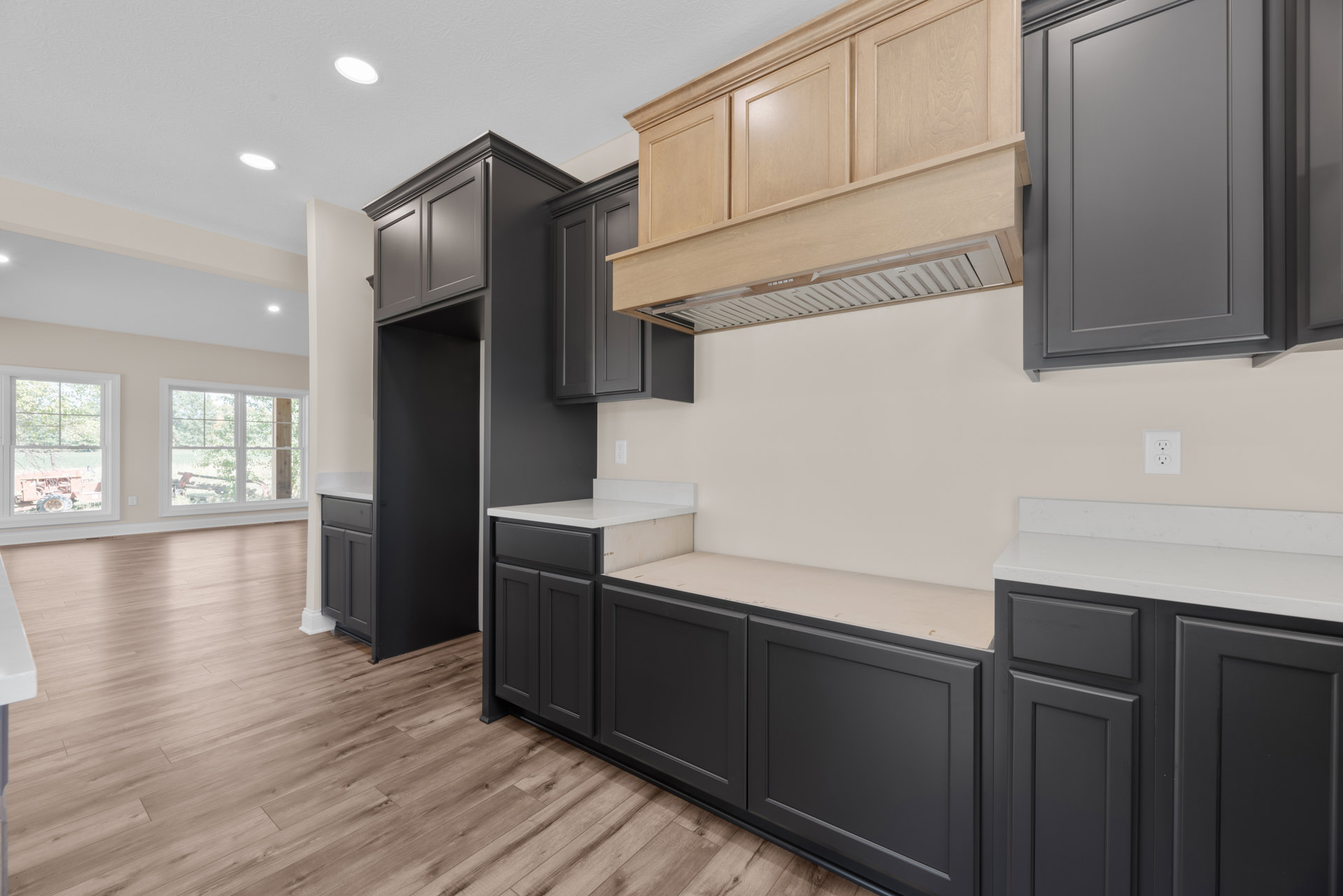 Black kitchen cabinets with matte finish, wood plank flooring, grey cabinet with wooden trim, white electrical outlet, windows showing tractor and tree views, close-up detail of