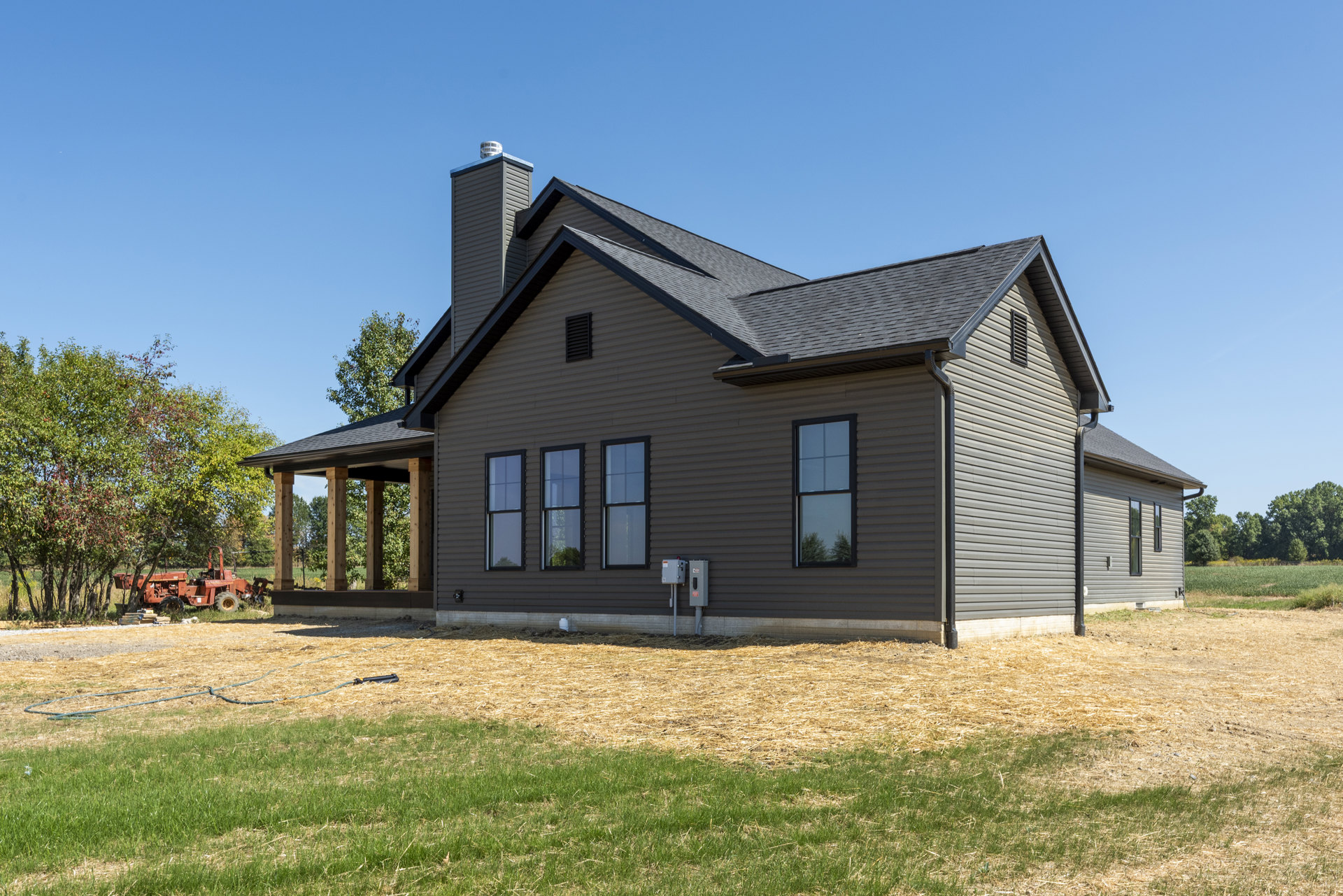 Modern cottage-style home with black-framed windows, surrounded by a green grass lawn, mature trees, and a tractor parked beside the house.