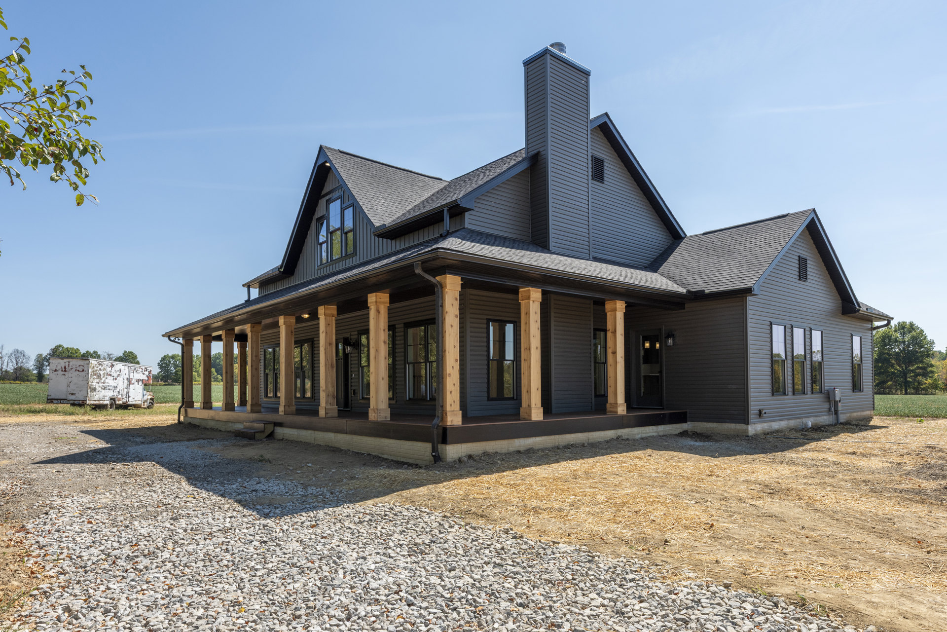 Wide front porch with white columns, gravel and grass yard, black-framed windows, tree branches with green leaves, white truck parked beside house
