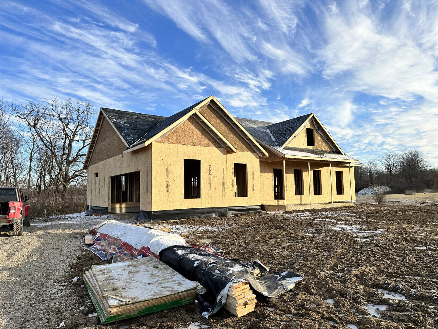 Framed house under construction with exposed wood beams, pile of lumber on dirt ground, blue sky overhead, partially installed windows