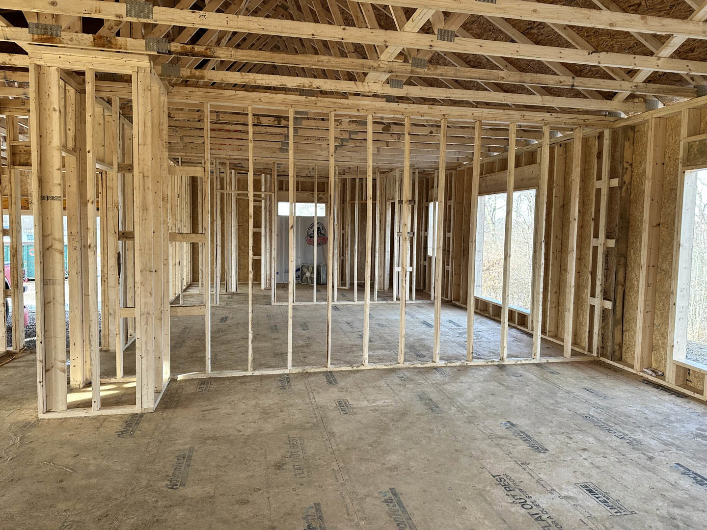 Open room with exposed wood ceiling beams, concrete floor marked with black and blue labels, white walls, large window overlooking trees.