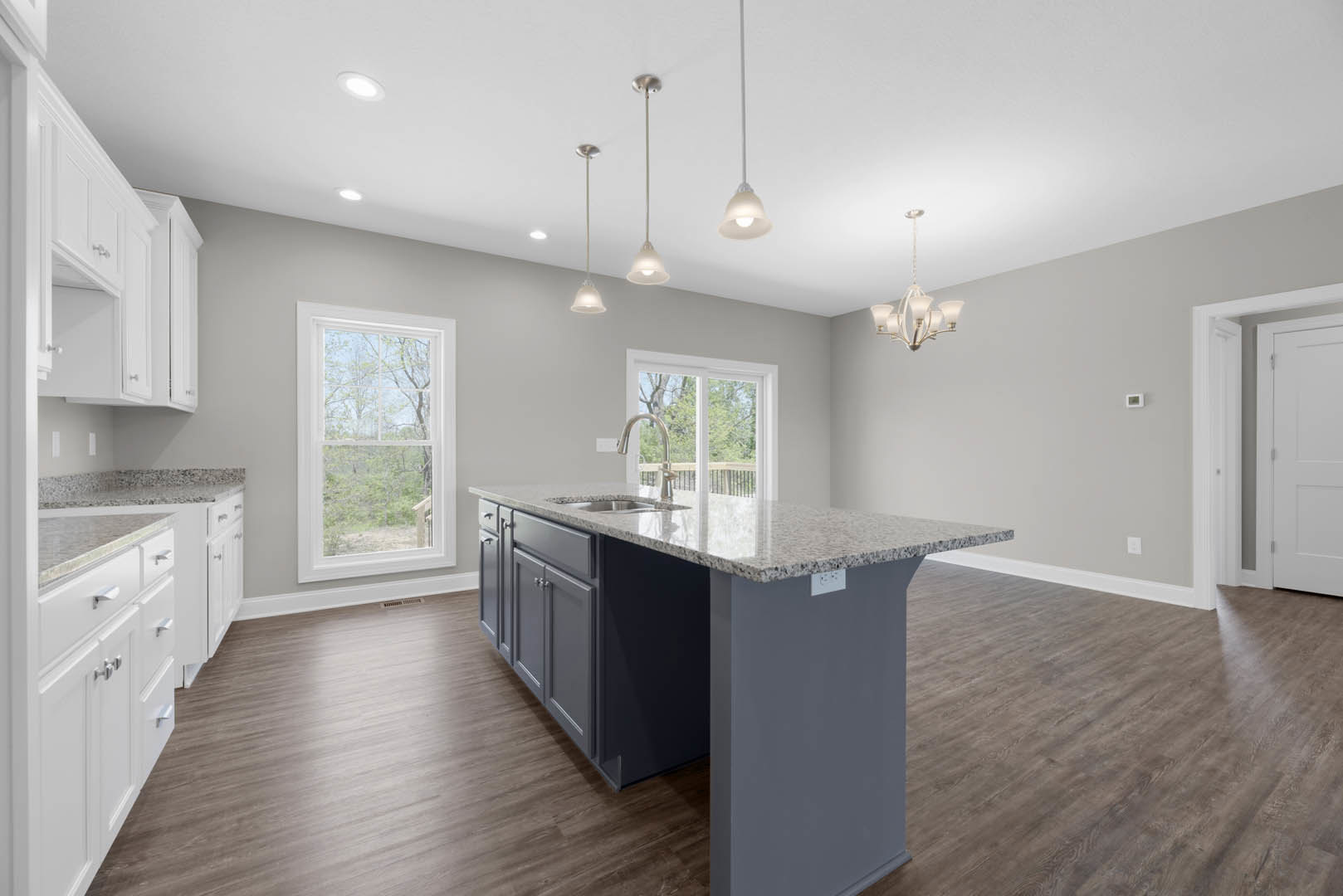 Open kitchen with wood flooring, central island featuring marble countertop and built-in sink, white cabinetry, window overlooking trees, staircase in background, white door with