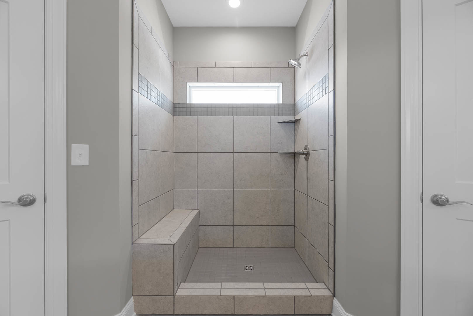 Tile shower with built-in bench, window above, chrome shower head, linear floor drain, white light switch on adjacent wall