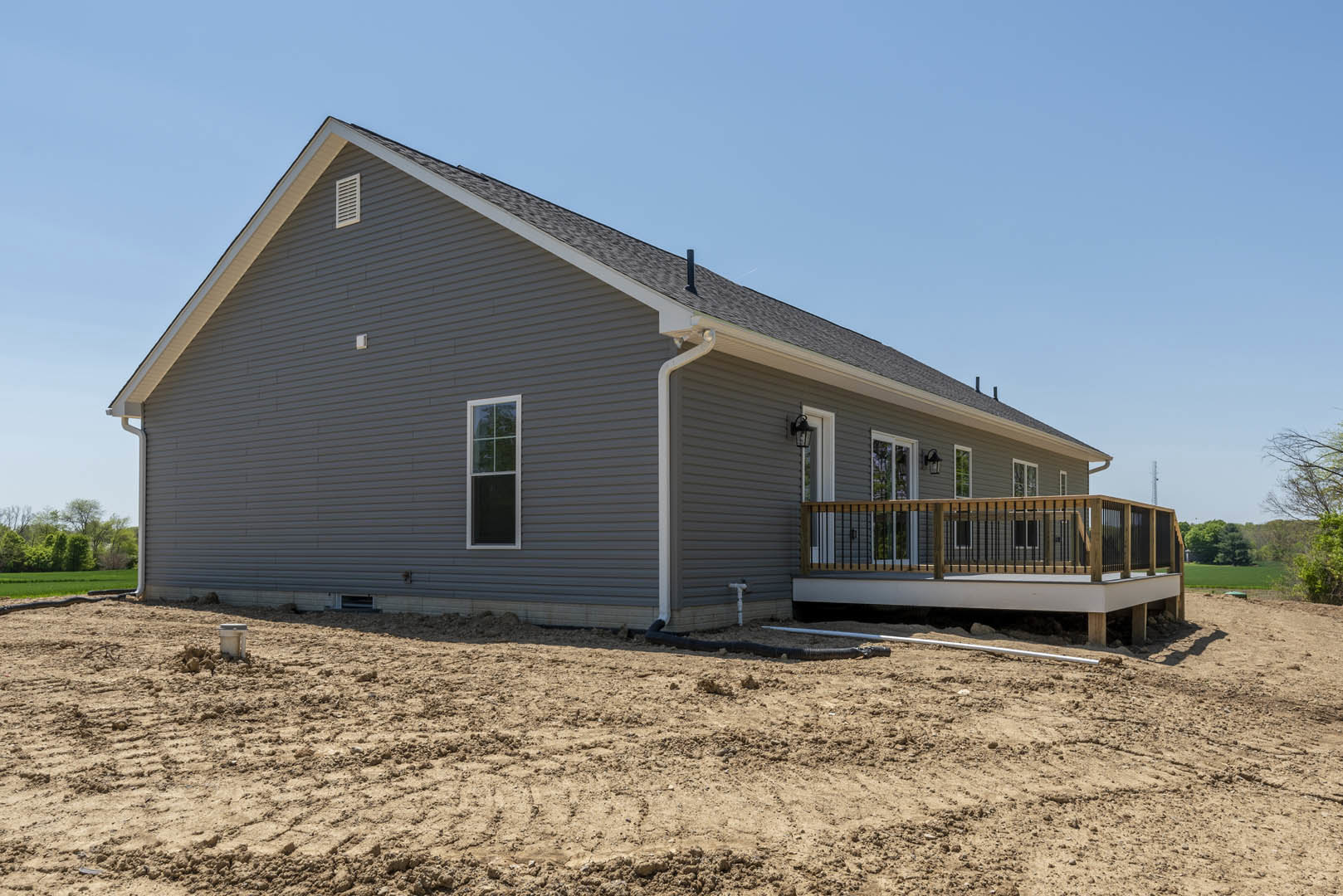 Partially built house with white-framed window, wooden deck featuring railing, covered porch, exposed dirt field with visible pipe, and cluster of trees in background