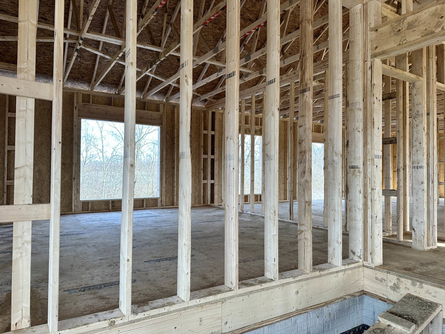 Room with concrete floor, large window, and wood-paneled wall; exposed wooden ceiling beams and unfinished construction details visible.