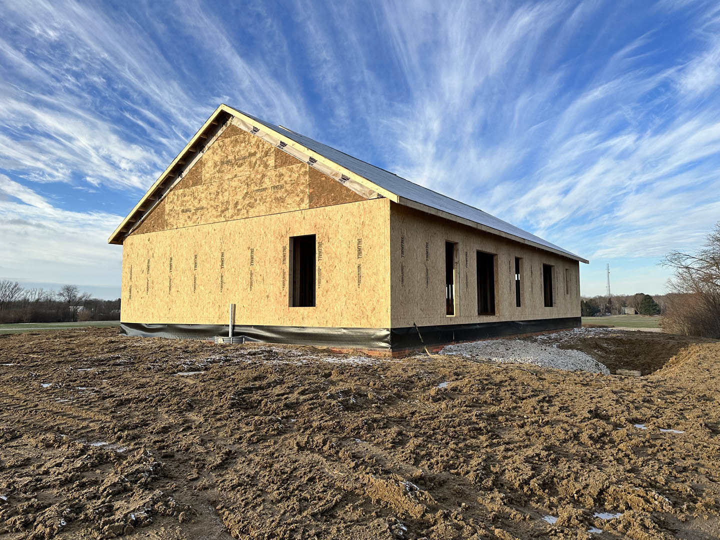 Wood-framed house under construction on dirt lot, unfinished windows, blue sky with scattered clouds, leafless trees in background