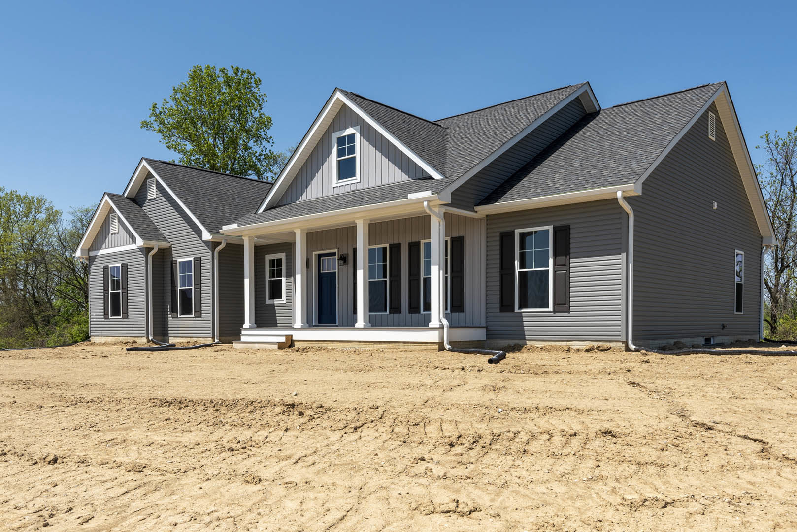 Wood-framed house under construction on bare dirt lot, white porch, white-framed windows, leafy tree nearby, blue sky, Robert Frost Farm visible in background