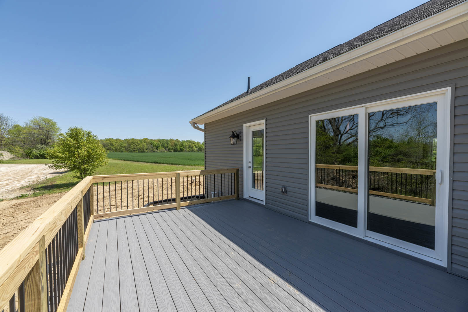 Wooden deck bordered by a horizontal slat fence, overlooking a grassy yard with mature green trees; house roof with vent pipe visible, sandbox area near deck, and close-up of