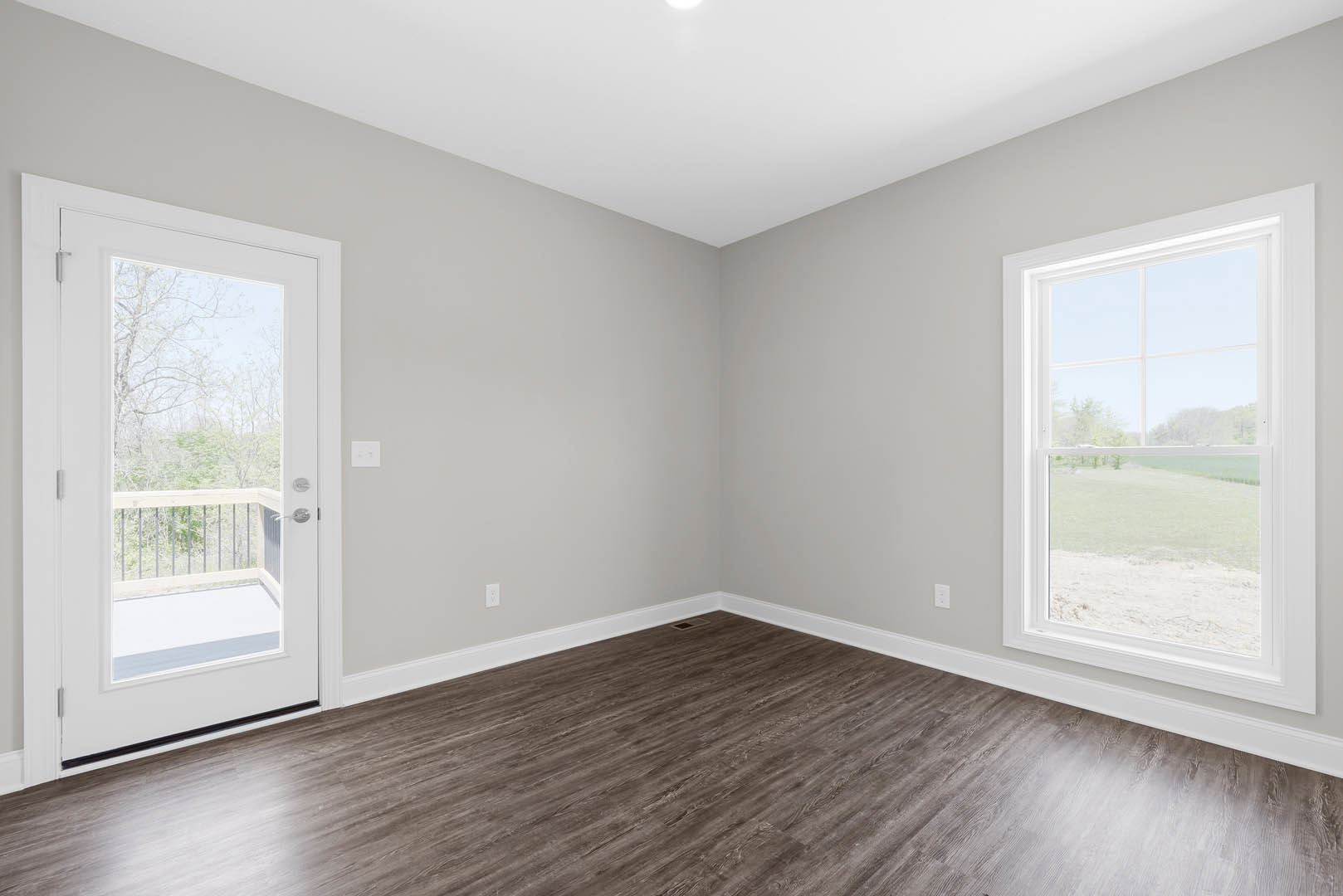 Wood-floored room with white baseboards, glass-paneled door, large windows overlooking a field, and metal railing visible near entry.