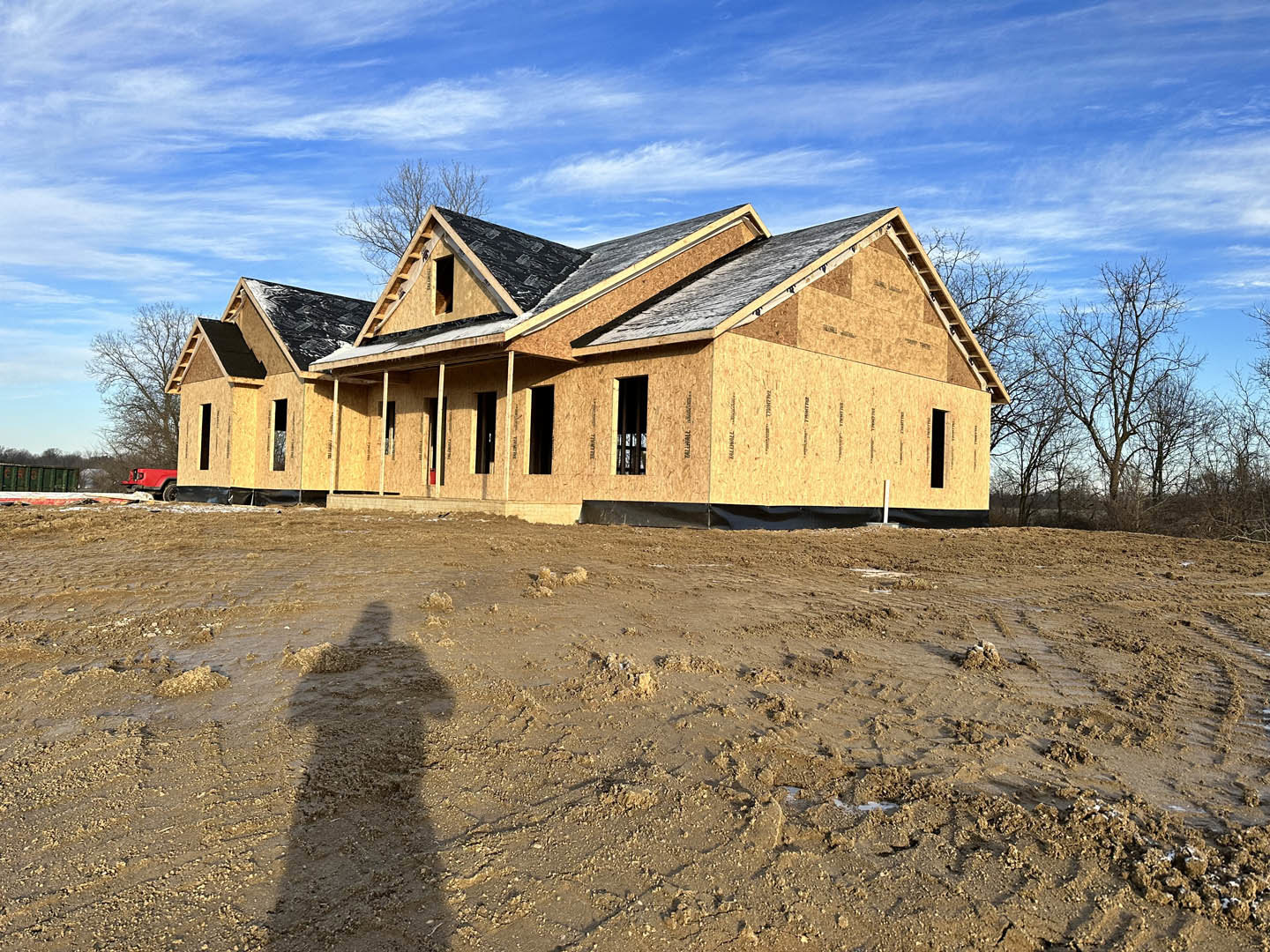 Framed house under construction with exposed roof, muddy ground, leafless tree, and clear blue sky; shadow of person walking across dirt foreground