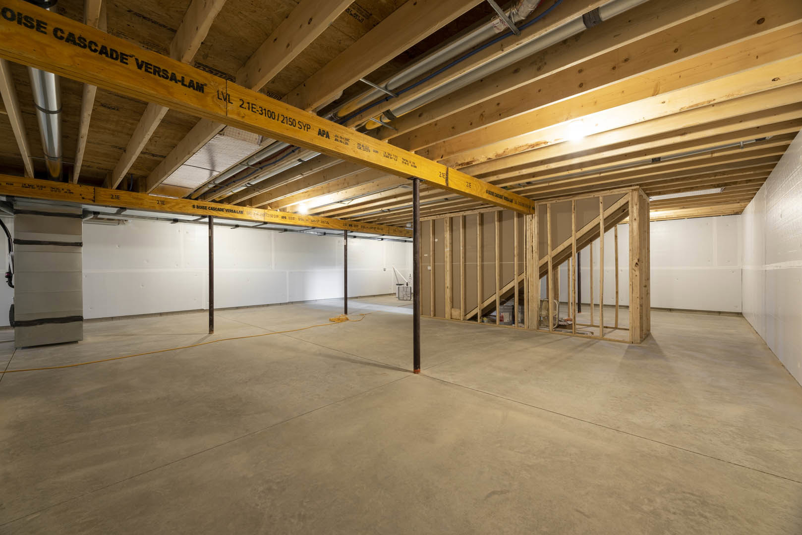 Exposed wooden ceiling beams with visible pipes, concrete floor featuring a basketball hoop, unfinished wooden framing, staircase, and a sign attached to a beam in a house under