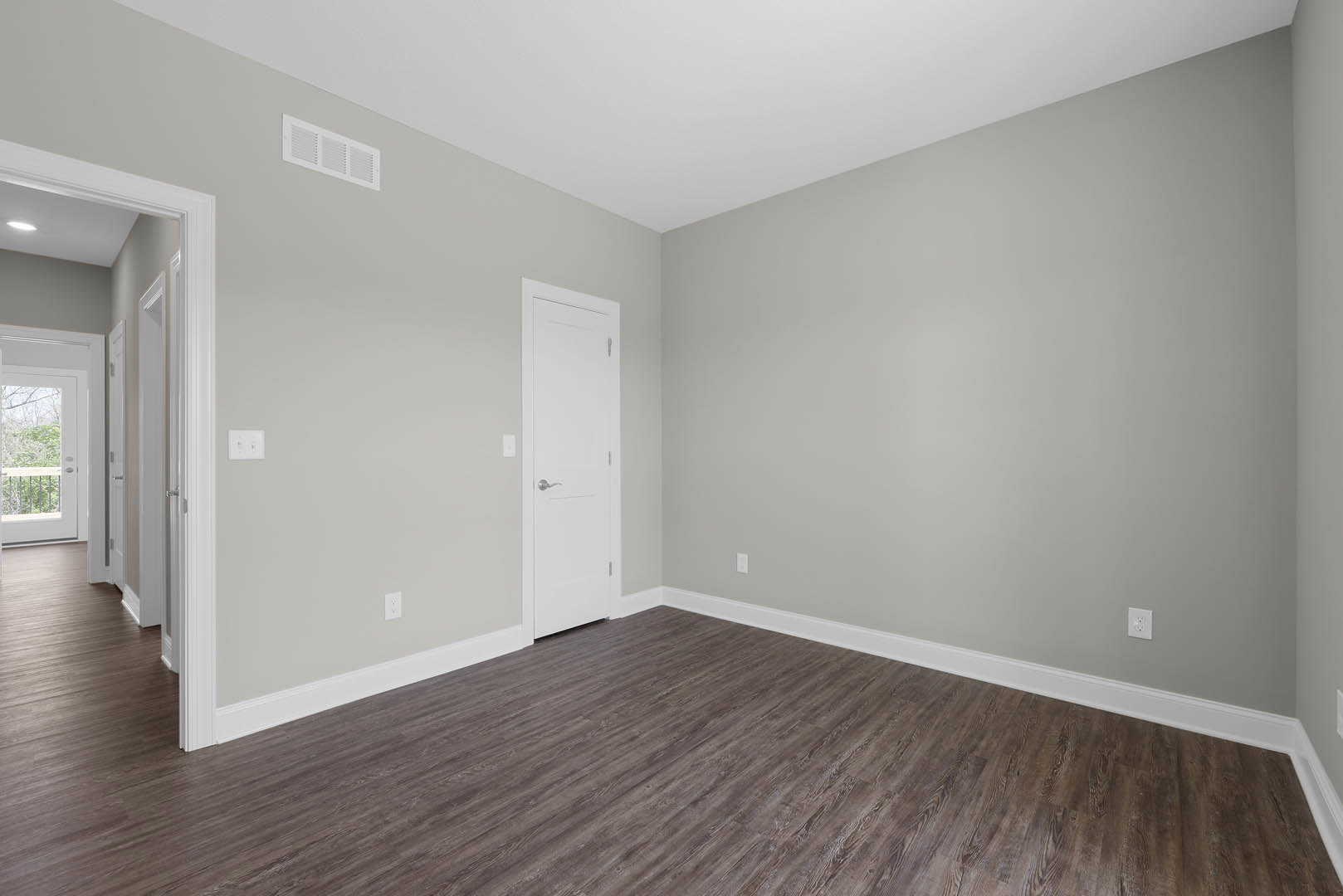 Room with light wood laminate flooring, white baseboards, white paneled door with silver handle, white ceiling vent, and a door with a window; neutral walls and modern finishes.