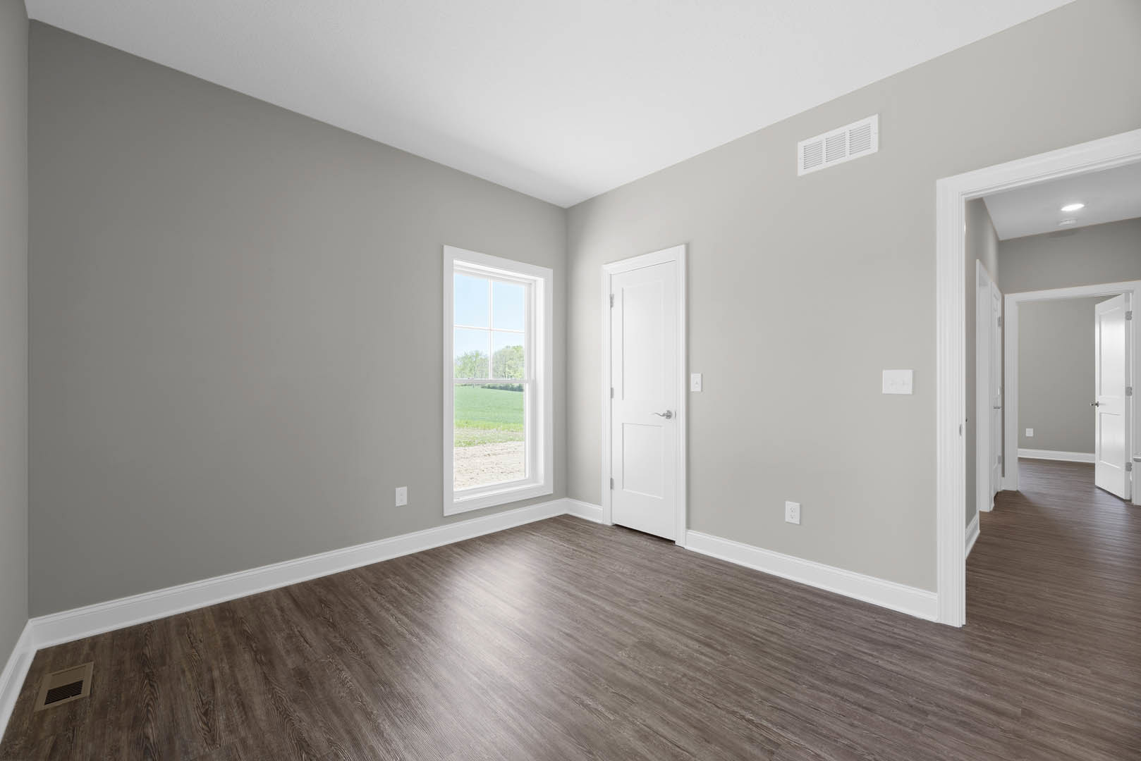 Bright room with laminate wood flooring, white plaster walls, a white door with silver handle, ceiling and wall vents, and a window overlooking a grassy field