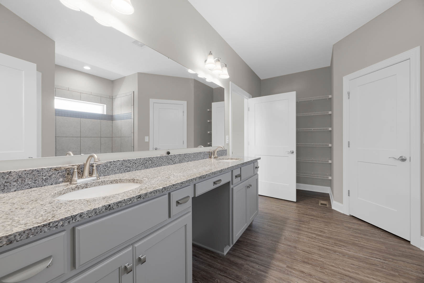 Bathroom with double white sinks on a stone countertop, wood plank flooring with floor vent, white door with silver handle, white shelf mounted on wall, cabinetry and drawers
