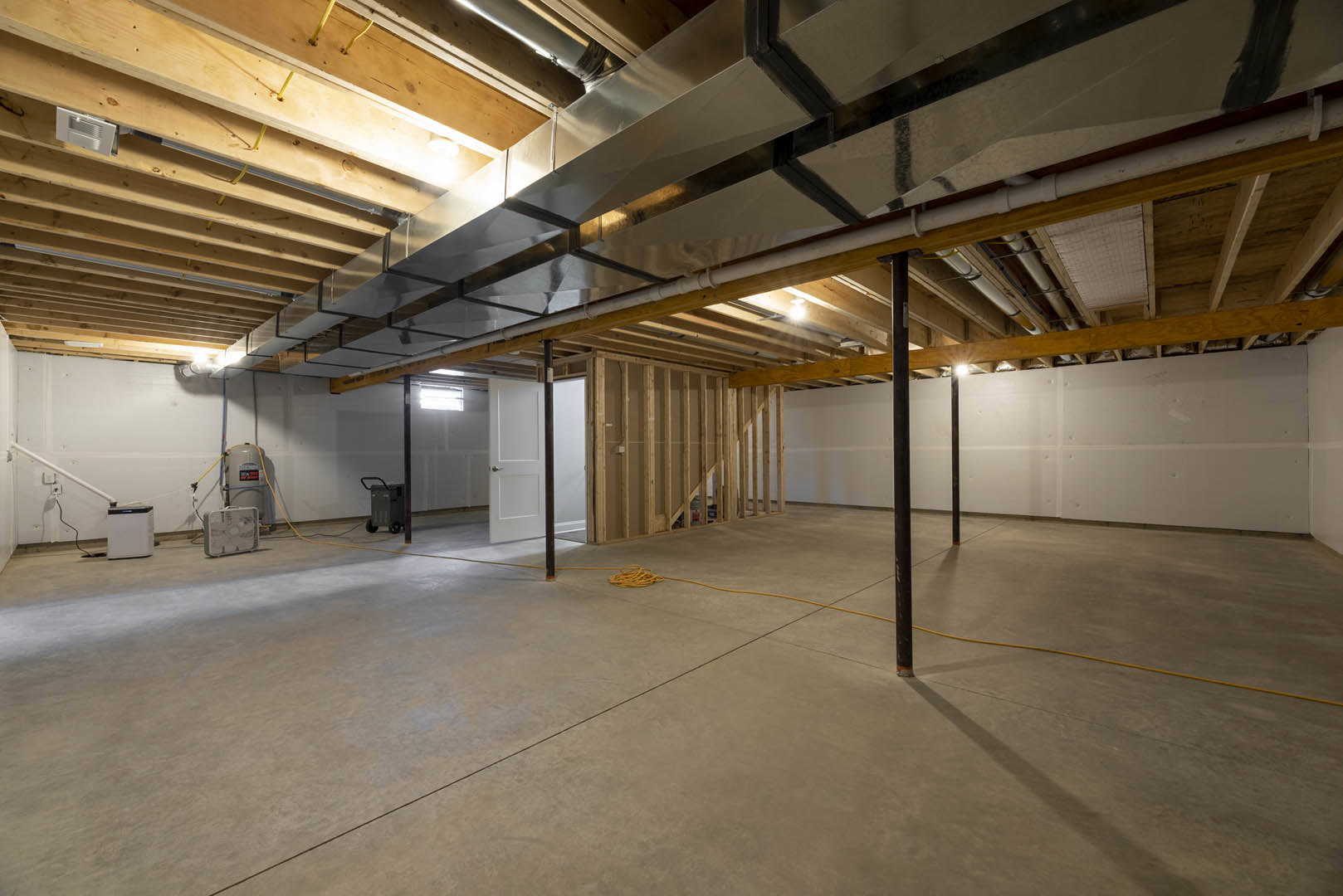 Concrete floor with yellow rope, metal ceiling with exposed pipes and beams, white door, water tank, white box with black top, white case with fan in an unfinished basement