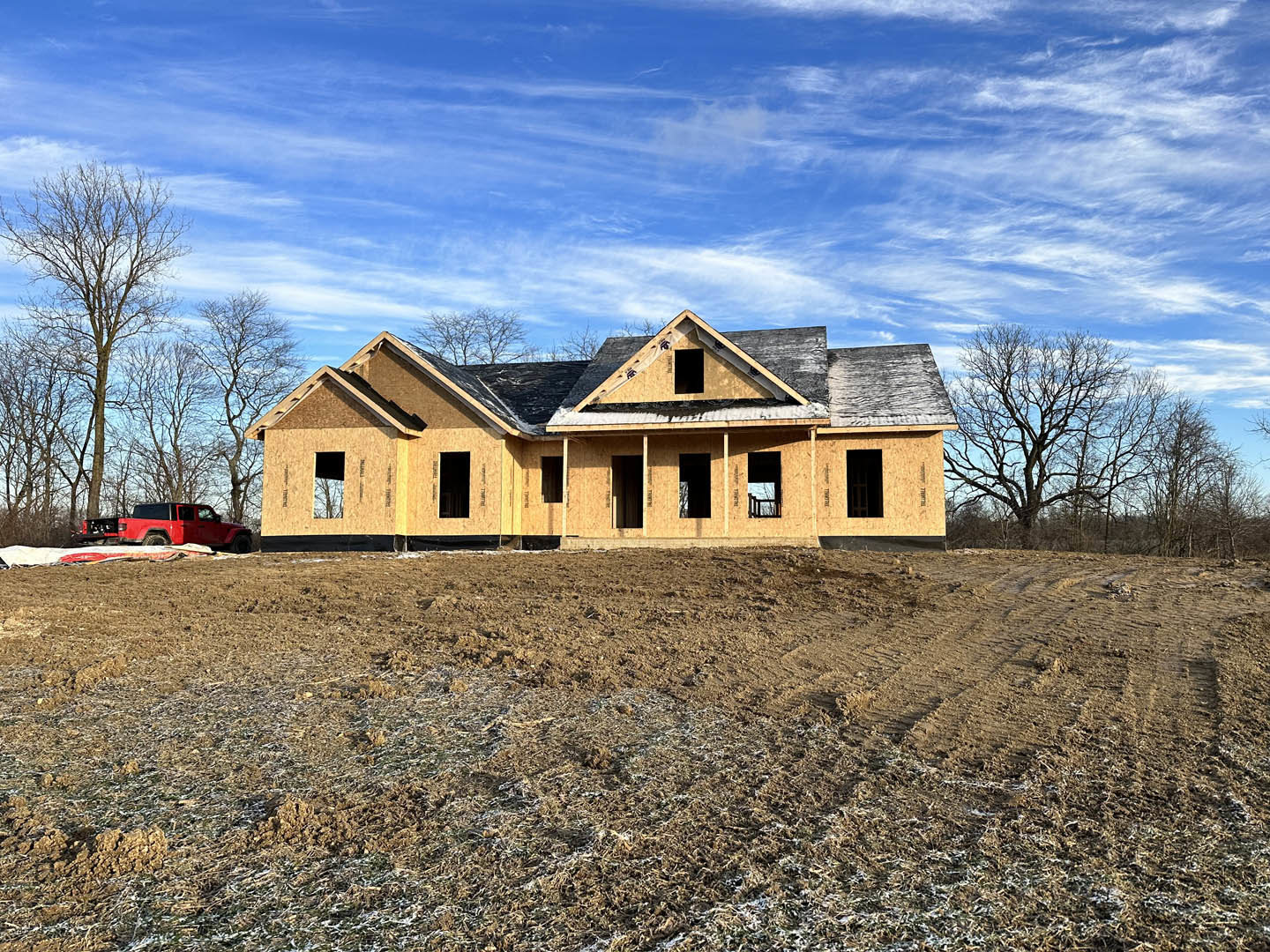 Partially built farmhouse with exposed framing, unfinished roof, and large dirt yard; red truck parked behind structure, leafless tree nearby, blue sky overhead