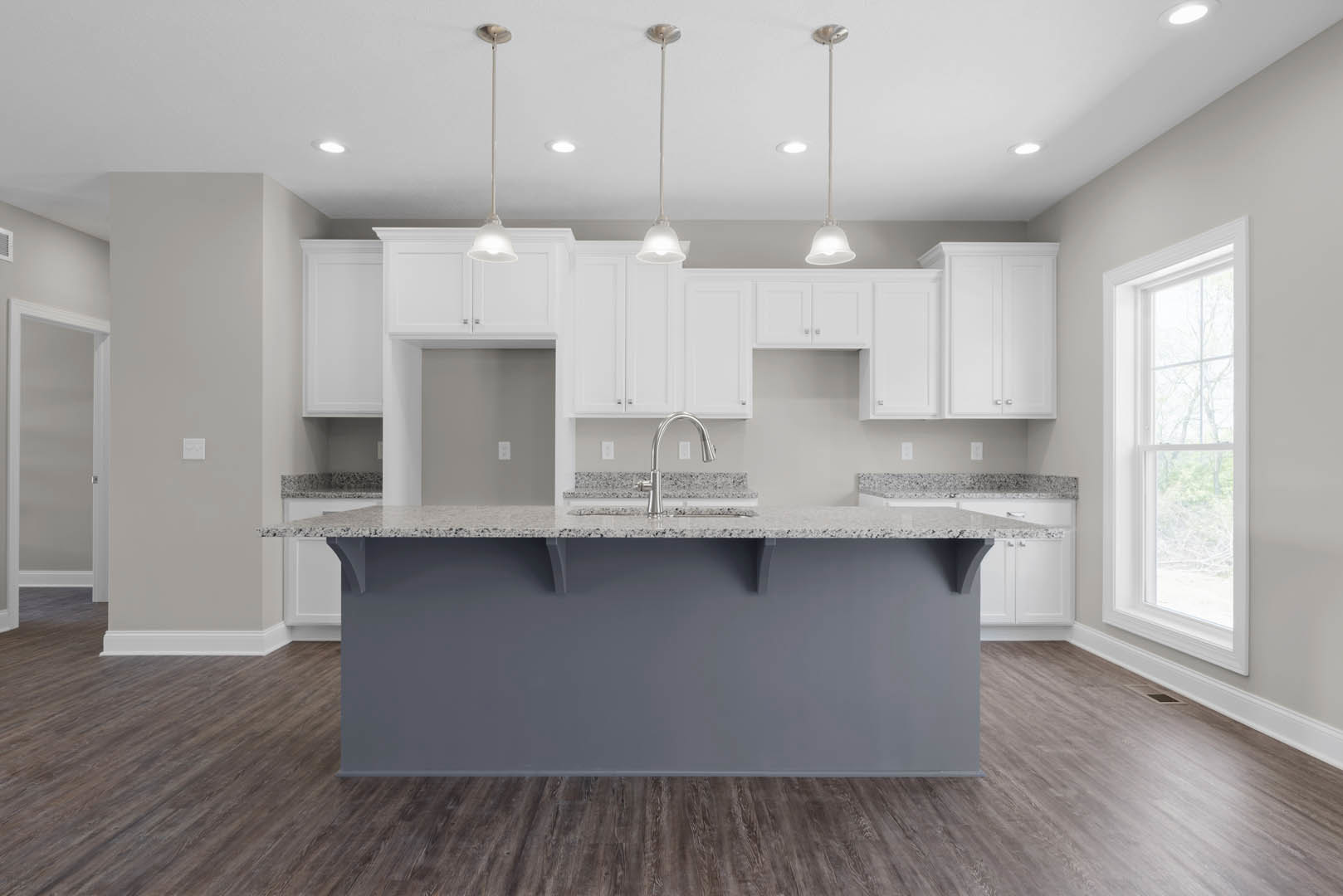 Gray kitchen island with quartz countertop, white cabinetry, stainless steel sink, tile backsplash, pendant light fixture, large window overlooking trees, hardwood flooring, white