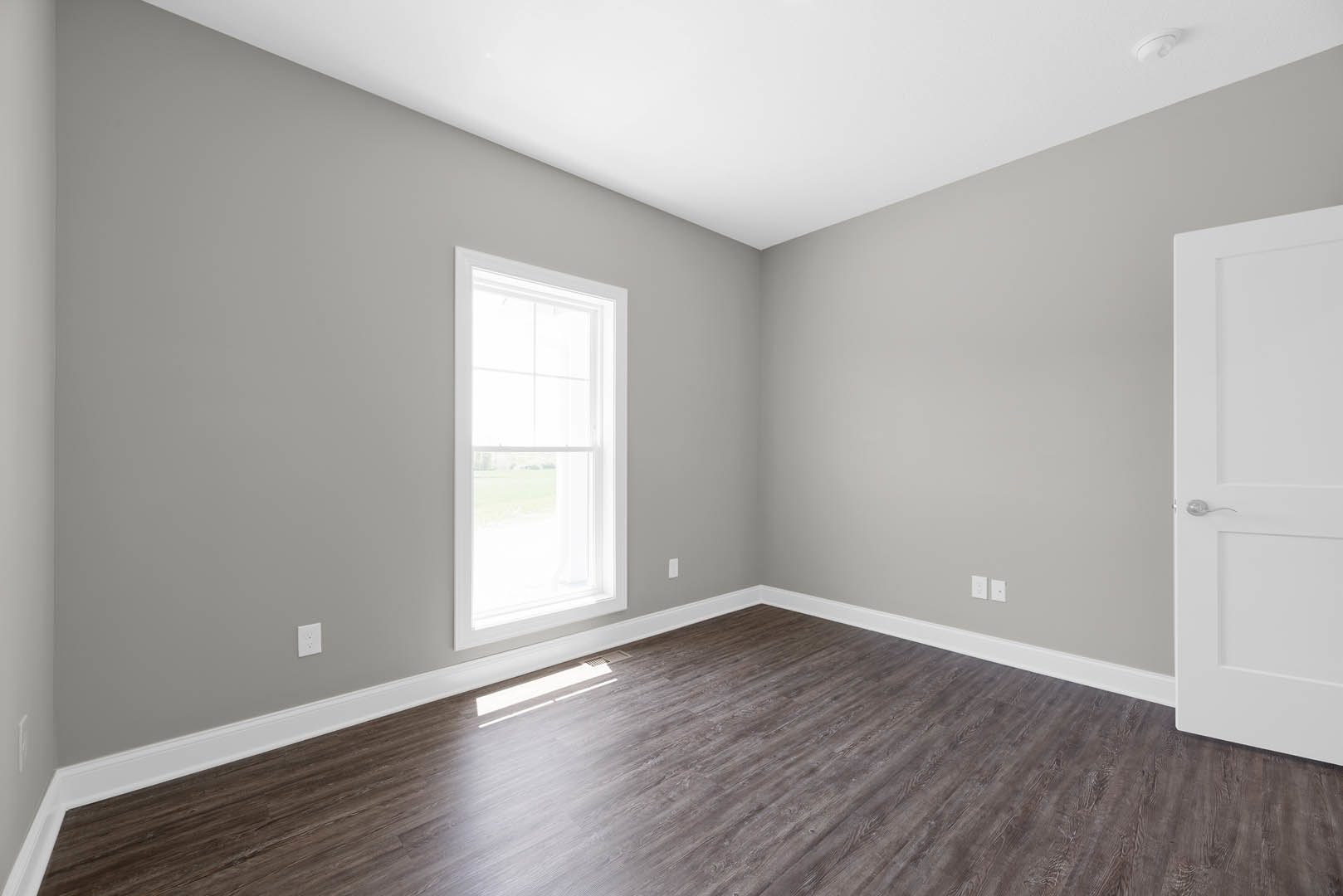 Sunlit room with wide-plank hardwood flooring, white walls, large window, white door, and smooth plaster ceiling.