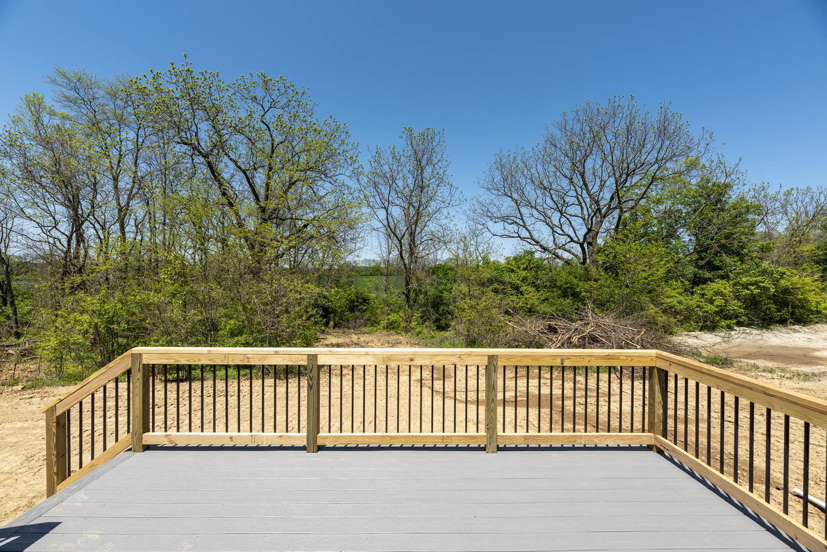 White-painted deck with wooden railing, bordered by a fence, surrounded by leafy green trees and one bare tree under a clear sky