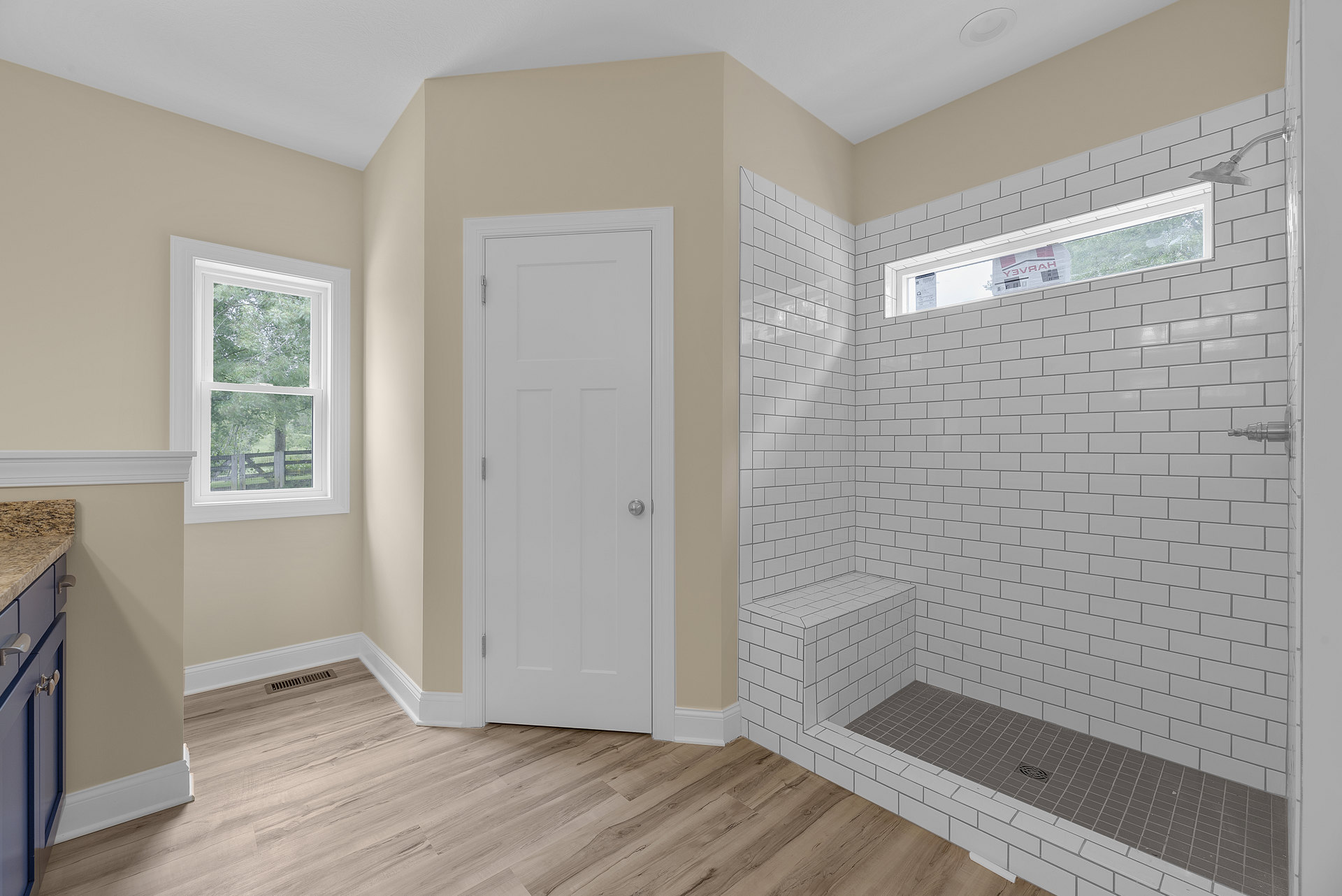 Bathroom with white tiled shower, window overlooking trees, white door with silver knob, tile floor with central drain, newspaper placed on window ledge