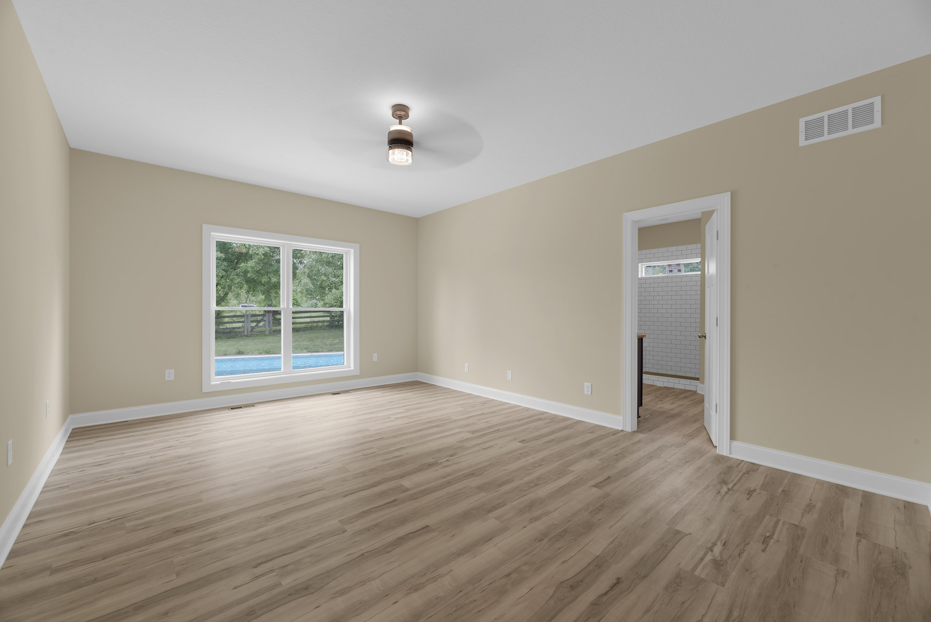Wood flooring in a bright room with a large window overlooking trees and grass, white plaster walls, ceiling vent, and a modern light fixture.