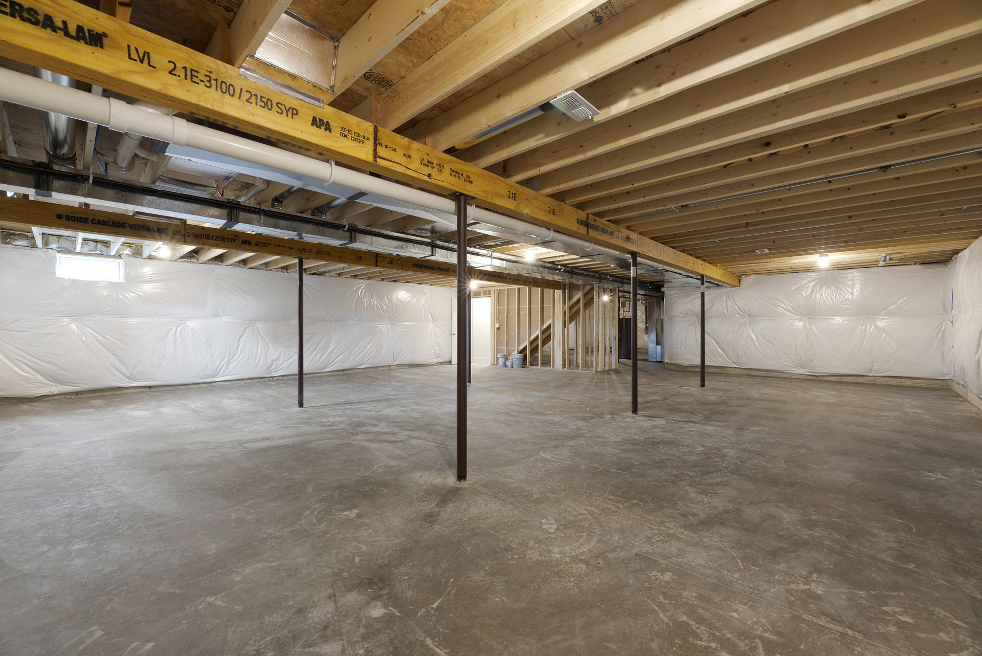 Basement room with exposed wooden ceiling beams, steel support pole, concrete floor, and white plastic tarp covering one wall.