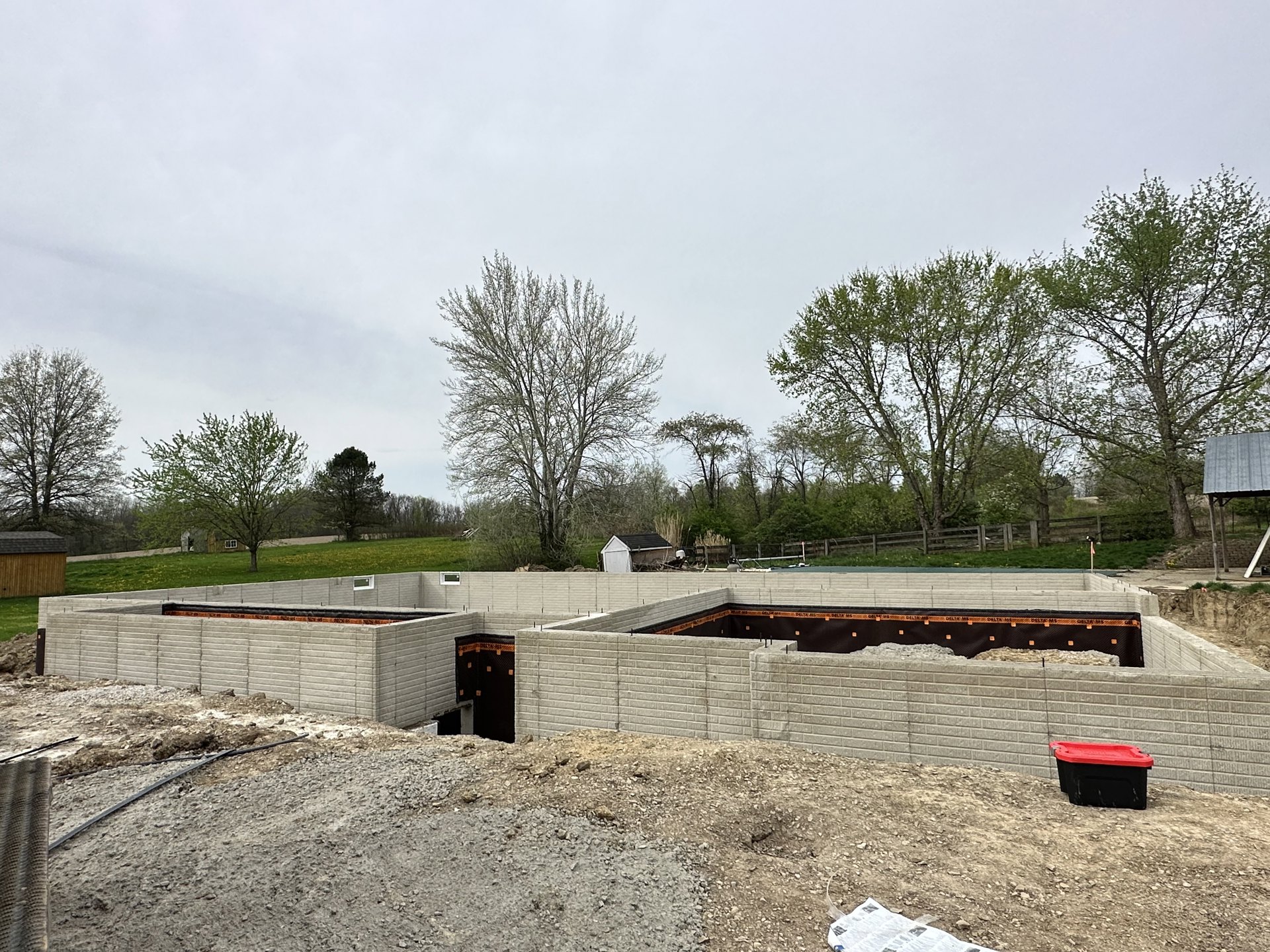 Grassy building site bordered by mature trees, partially constructed brick wall, black and red storage container, leafless tree, small shed with black roof under cloudy sky
