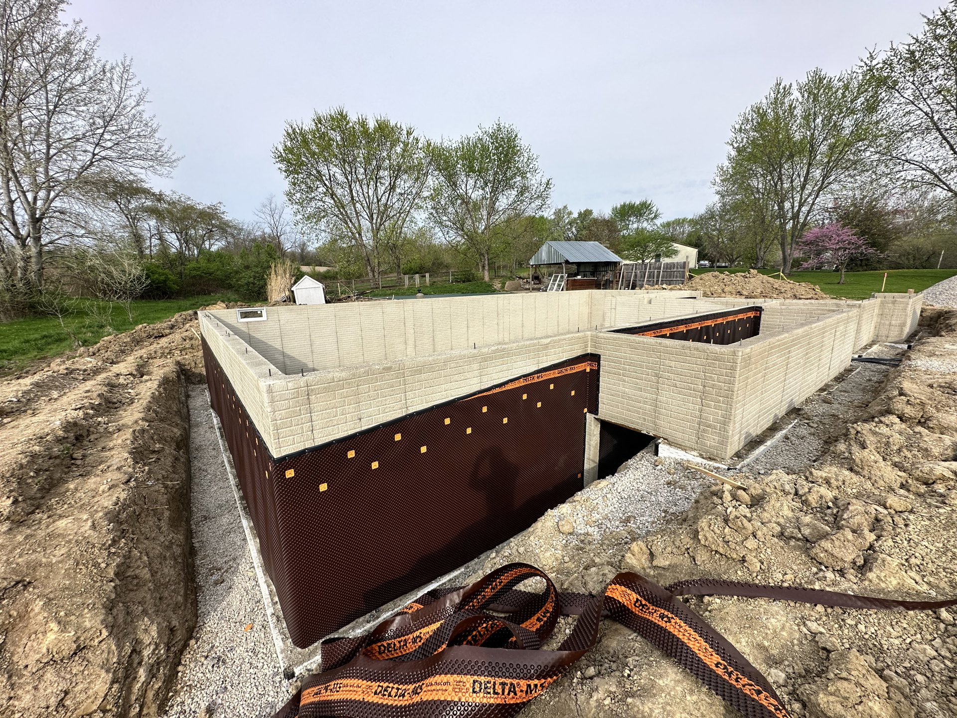 Partially built home with exposed foundation, open excavation, brick walls, unfinished roof, leafless tree nearby, and construction materials scattered on rocky ground