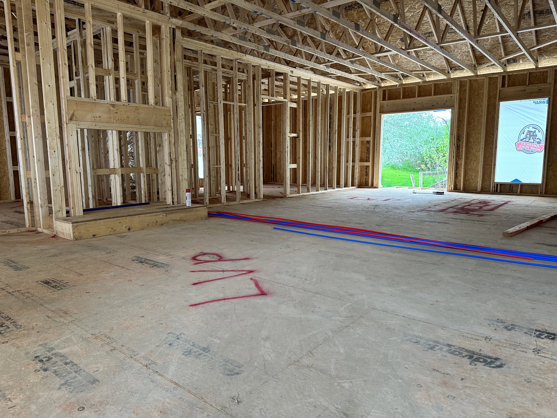 Unfinished room with exposed wood framing, concrete floor marked with red writing, red and blue plumbing pipes, large window overlooking fence and trees