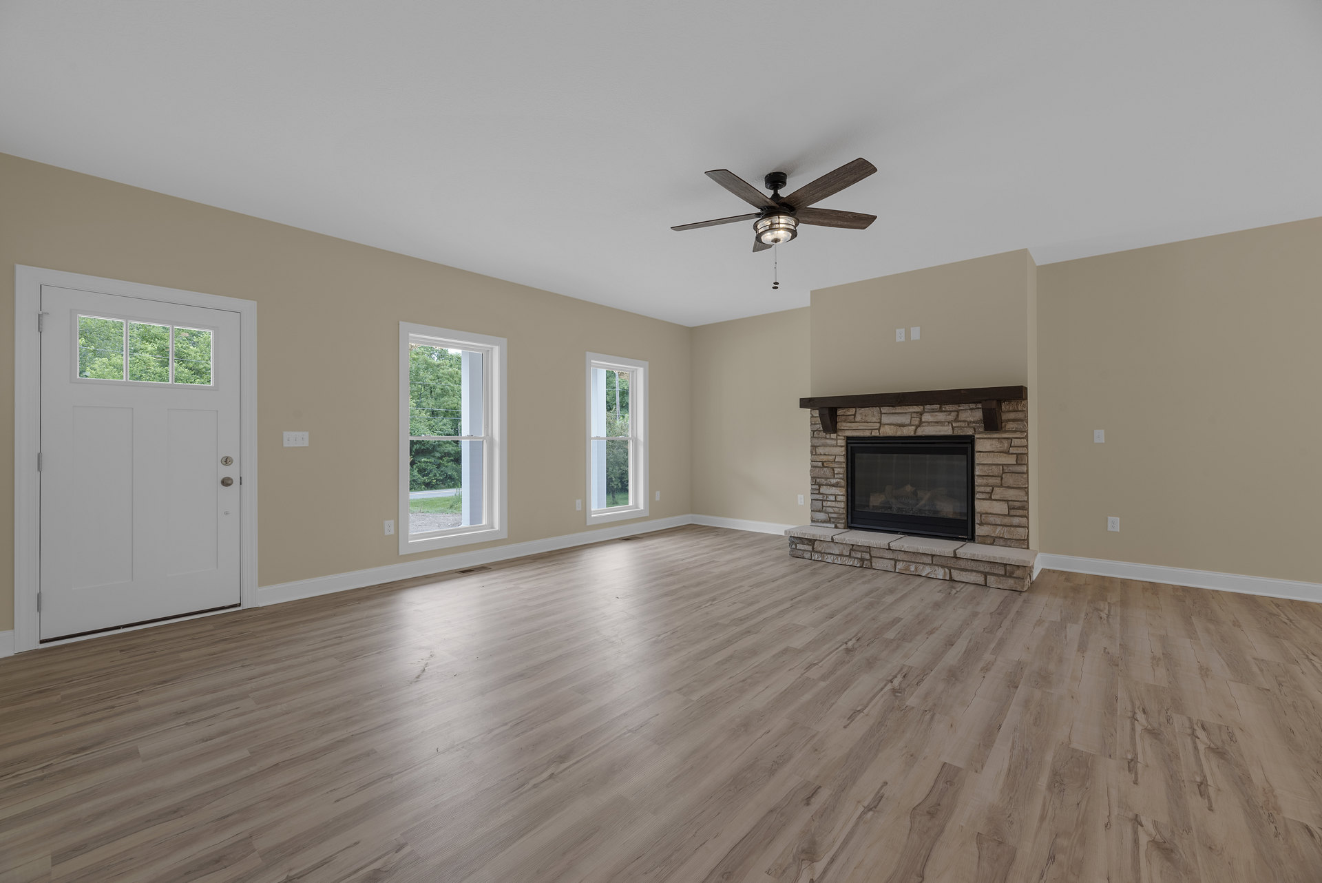 Living room with wood flooring, plaster walls, ceiling fan with light, glass door fireplace burning wood, large window showing trees, white door with window panel