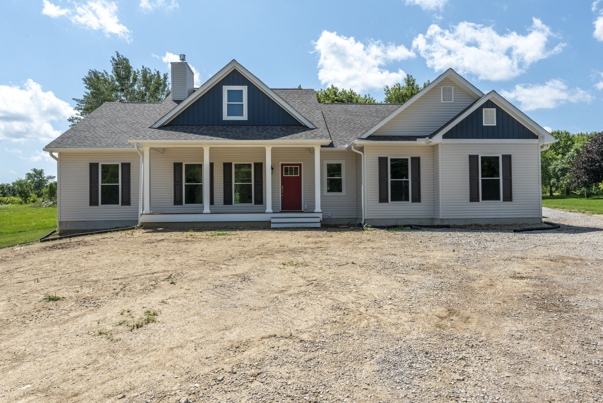 Two-story house with white siding, black shuttered windows, and a red front door, set on a dirt yard with a small porch and scattered trees under a cloudy sky