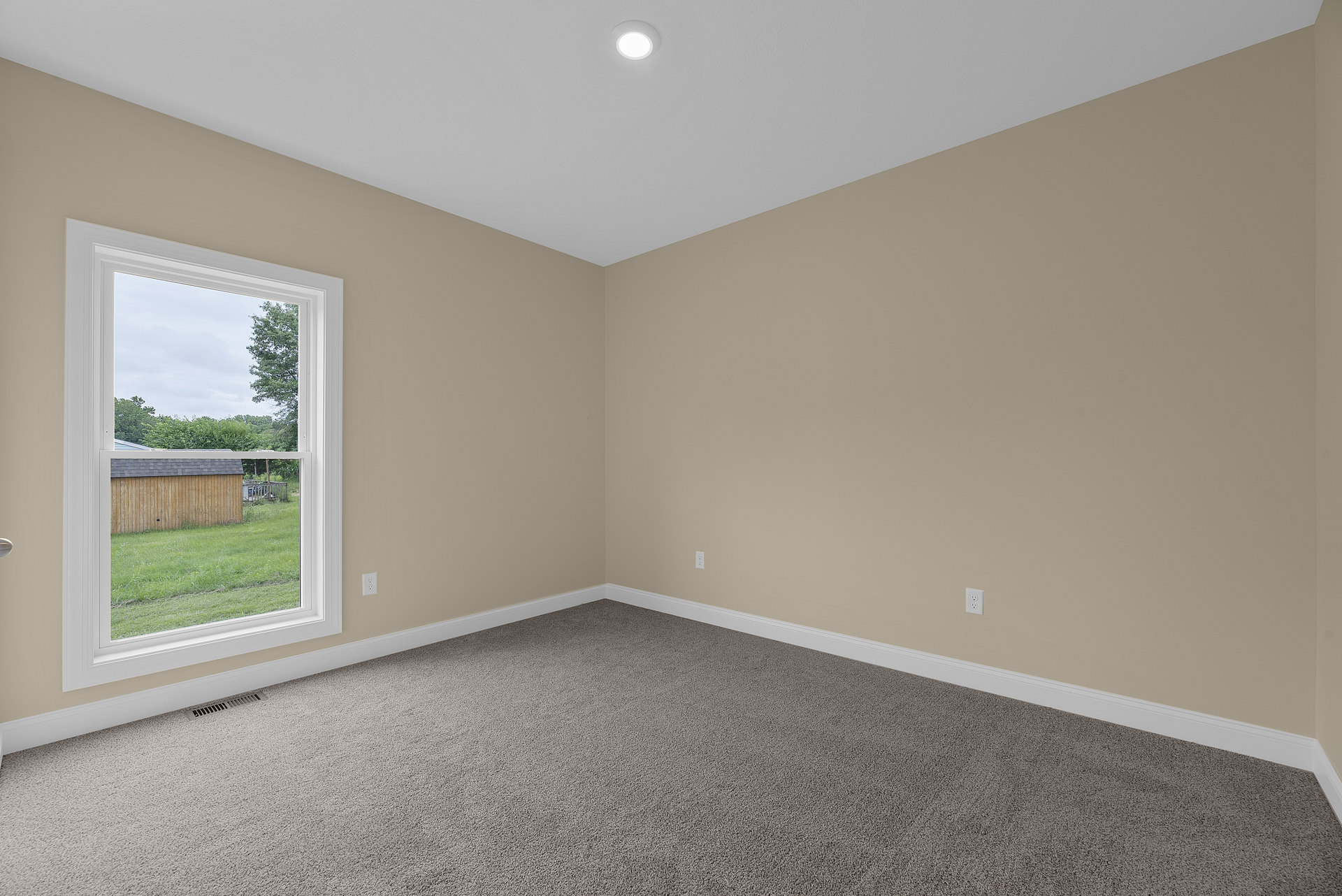 Bedroom with beige carpet, white walls, large window overlooking fenced backyard with shingle roof, recessed ceiling light, simple baseboard molding