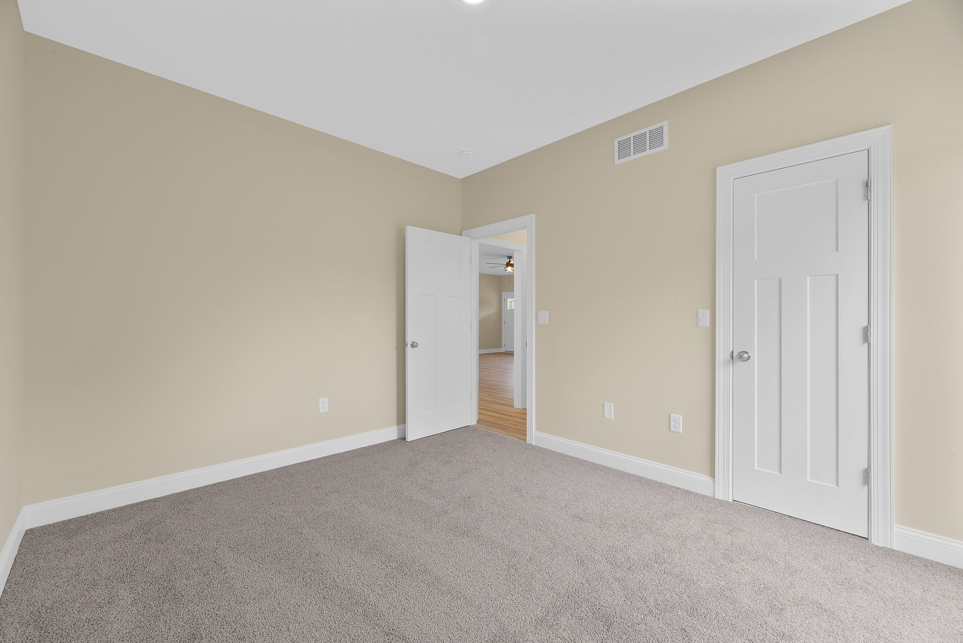 Beige-walled room with white doors featuring silver knobs, carpeted floor, wall vent, and white baseboards