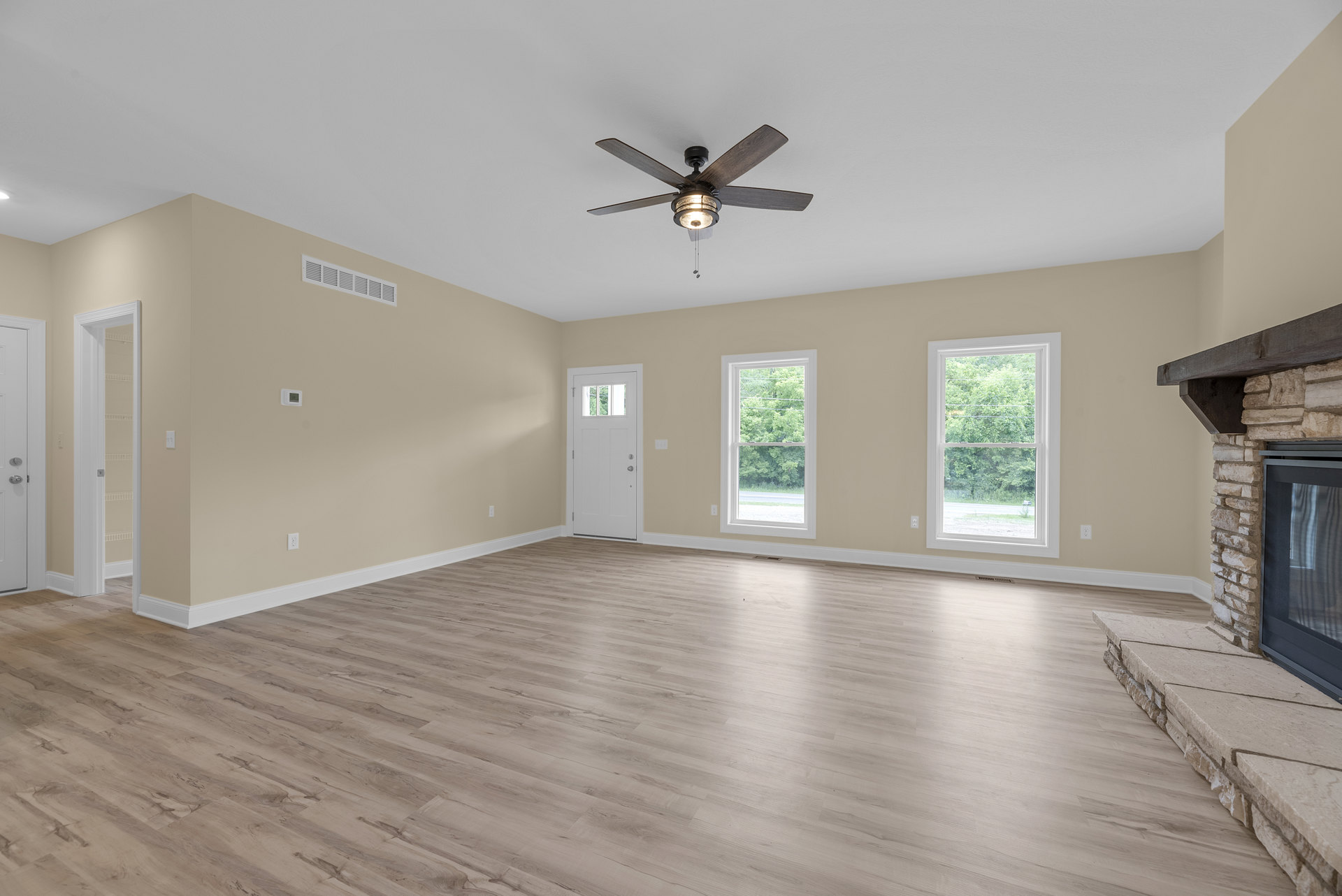 Ceiling fan with light fixture, wood flooring, white walls, window showing trees outside, white door with glass panel, fireplace partially visible