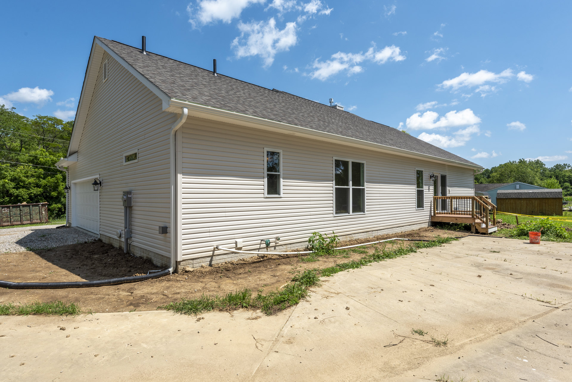 White siding house with large window, wooden deck featuring metal railings, concrete walkway bordered by green grass, garden hose coiled near foundation, gray shingled roof, trees