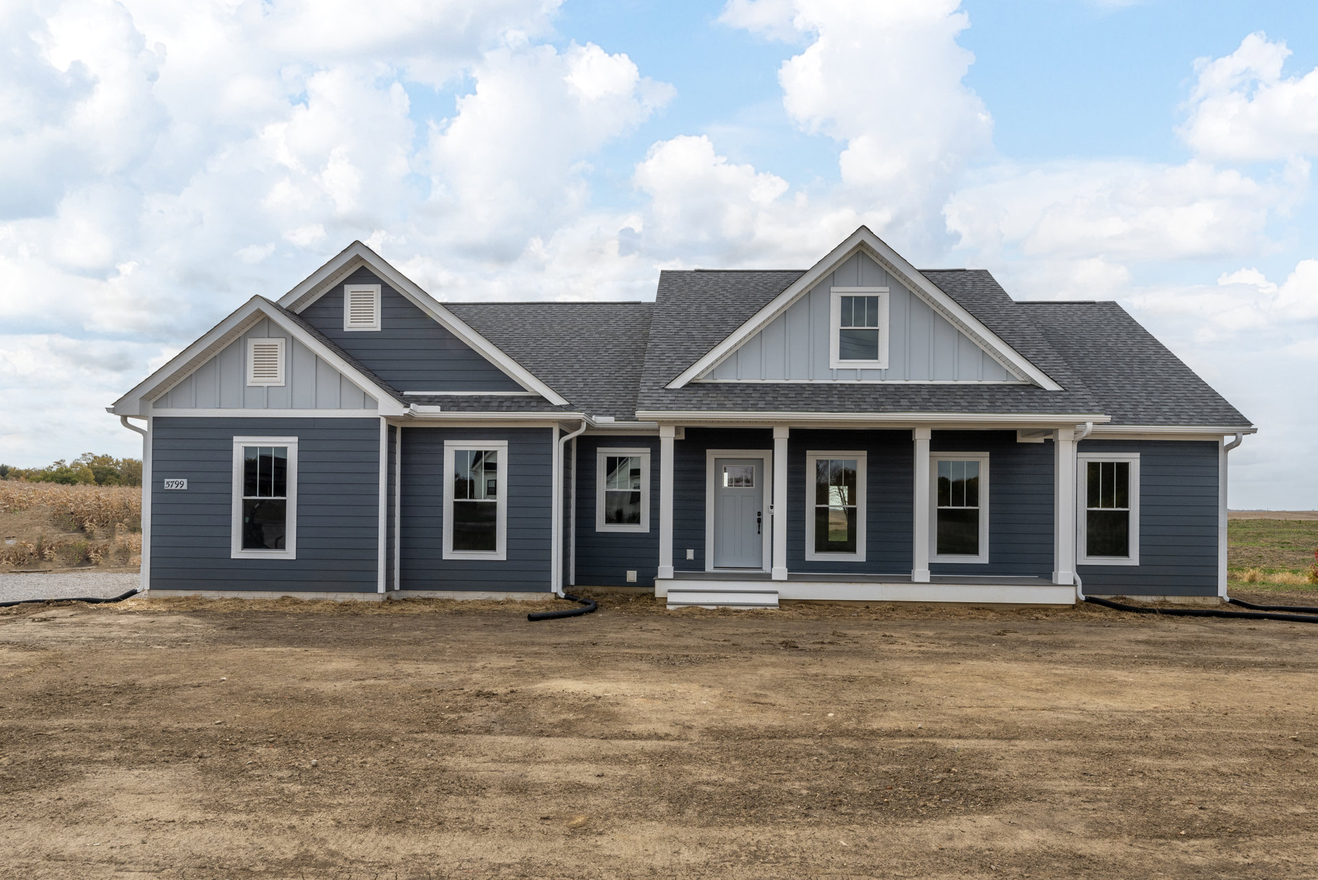 Partially built house with white siding, grey door, white-framed windows, and dirt field in foreground under cloudy sky