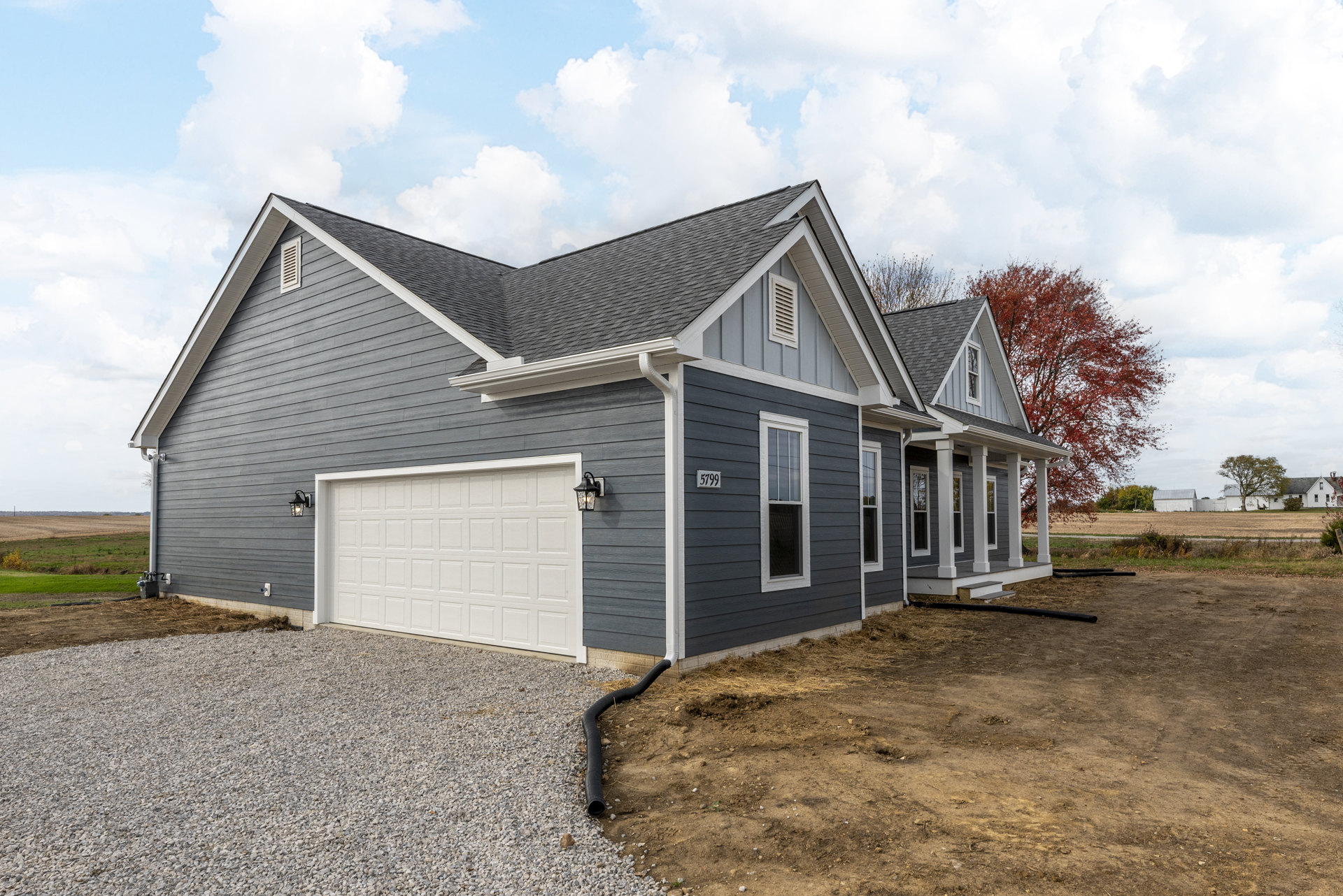 Grey siding house with white-framed window, white garage door, gravel driveway, red-leaved tree, and dirt ground.