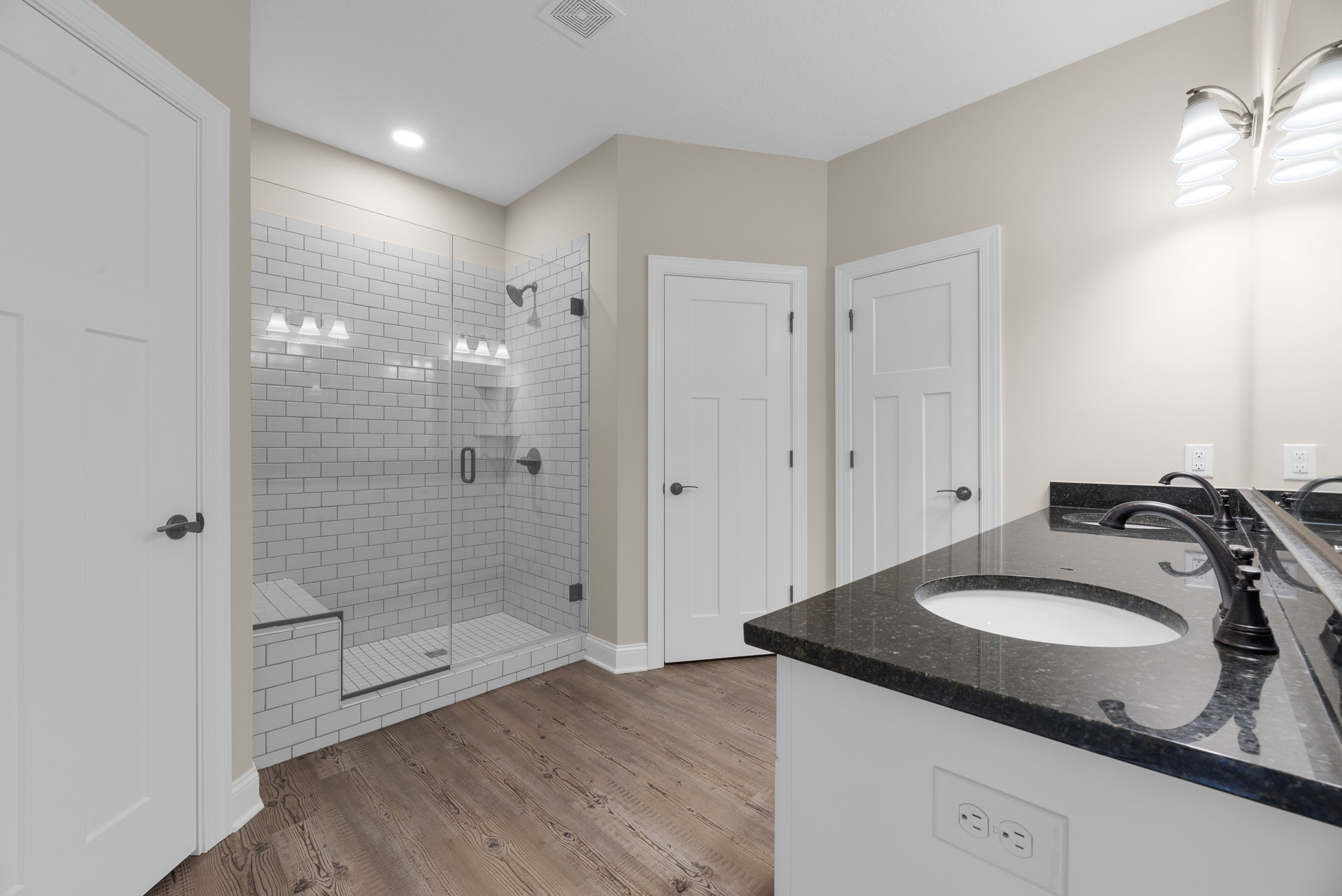 Bathroom with glass shower enclosure, white sink and faucet, light tile flooring, white door with black handle, neutral walls, modern cabinetry, and stone countertop.