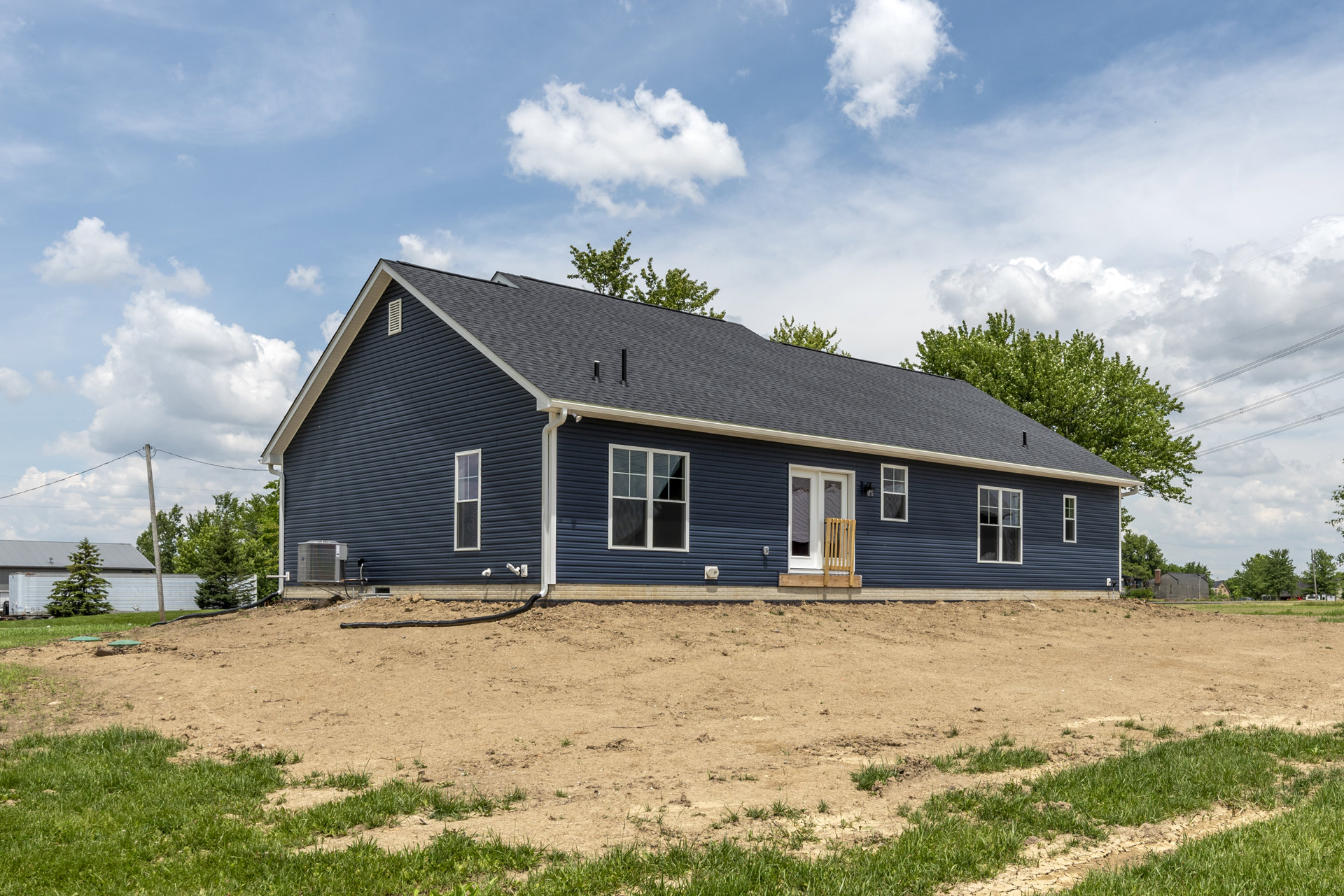 Blue house with white trim and covered porch, dirt field in foreground, trees and cloudy sky in background, wooden chair on window sill.