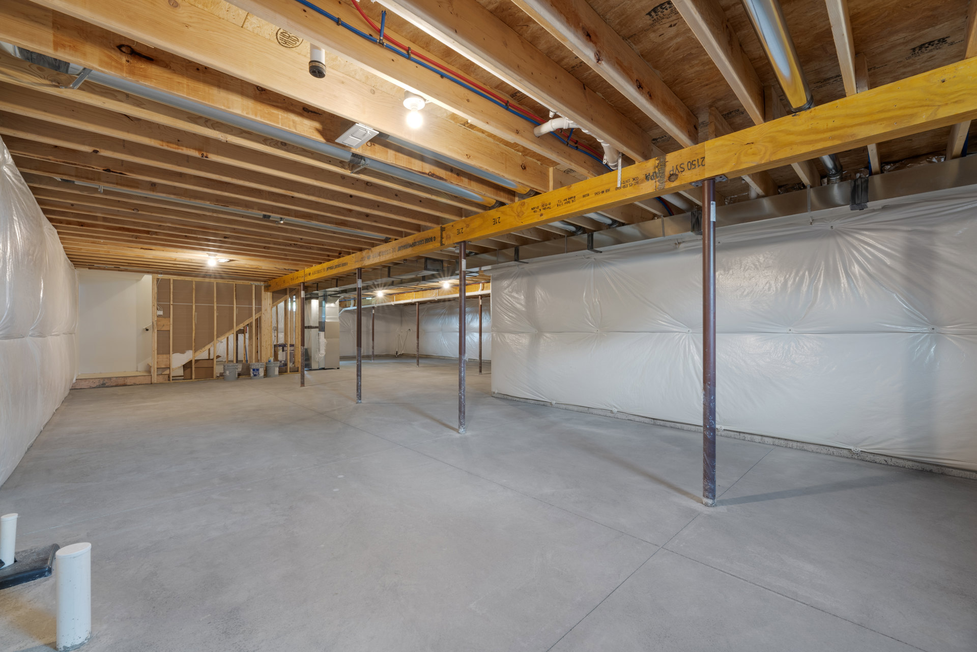 White walls and exposed wooden ceiling beams above a concrete floor with steel support poles, basement interior with composite insulation materials visible.