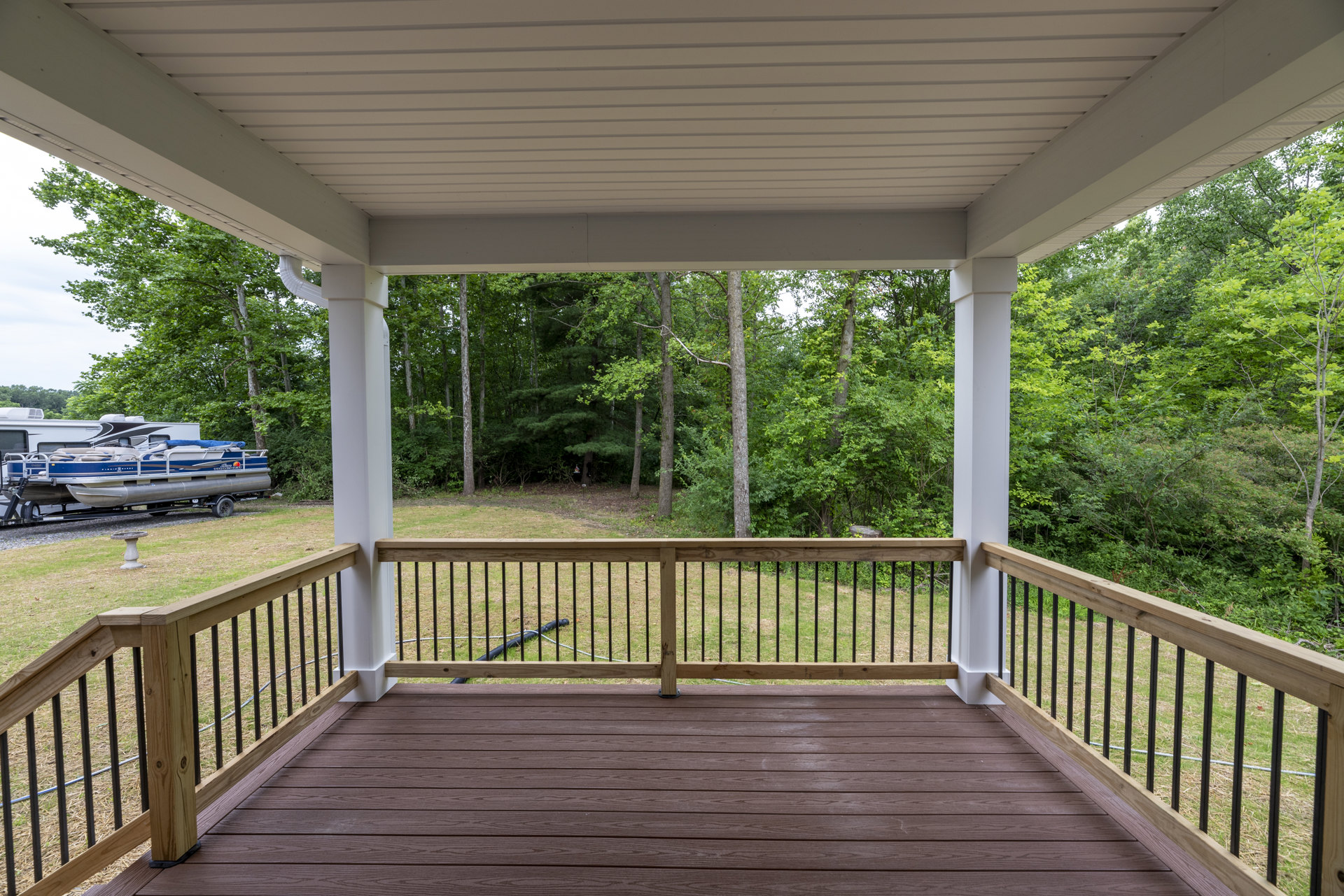 Wood deck with black metal railing, hose coiled near the wall, fenced yard, mature trees in background, boat on trailer parked nearby.