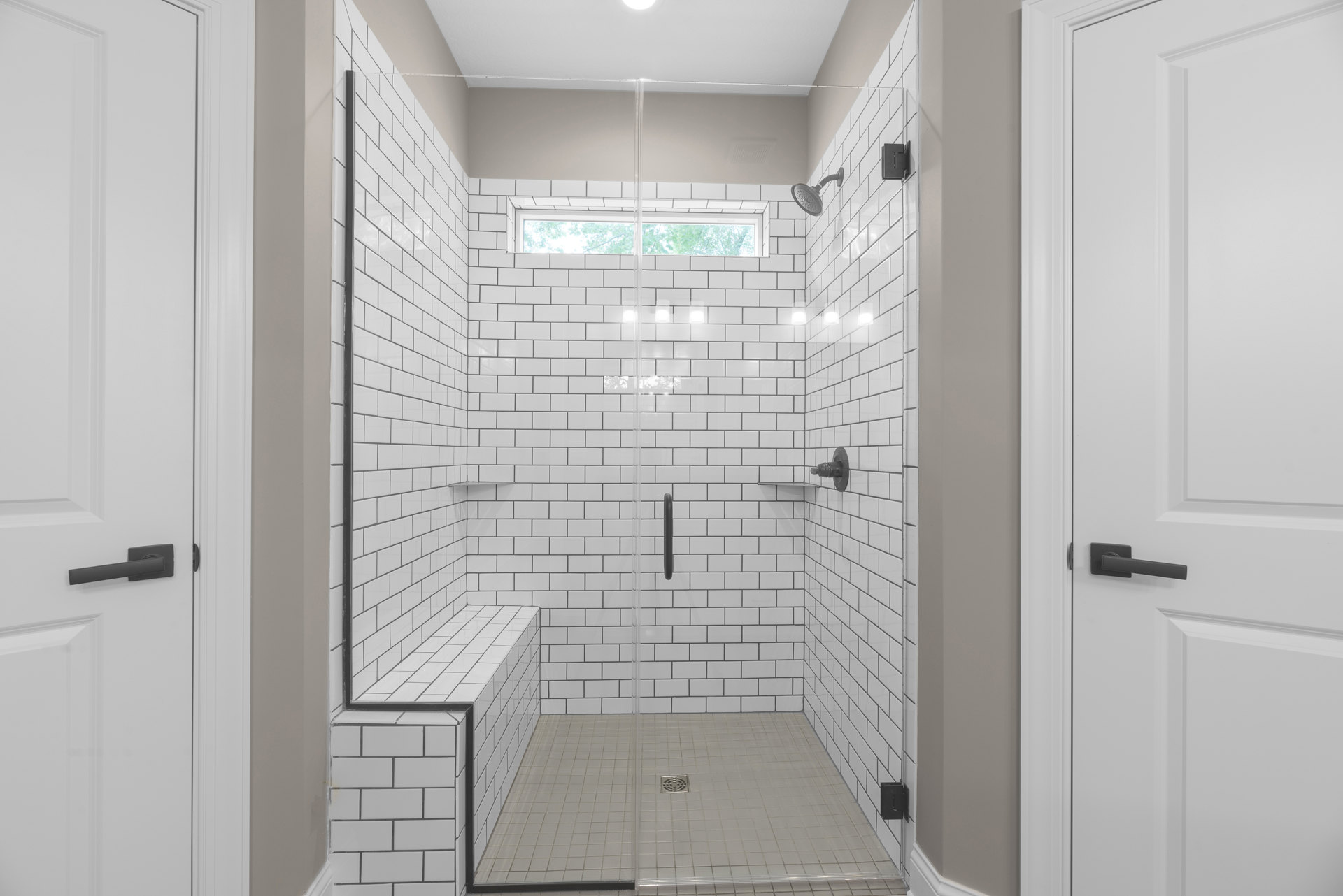Walk-in shower with clear glass door, white tile walls, built-in window, black hardware, and floor drain