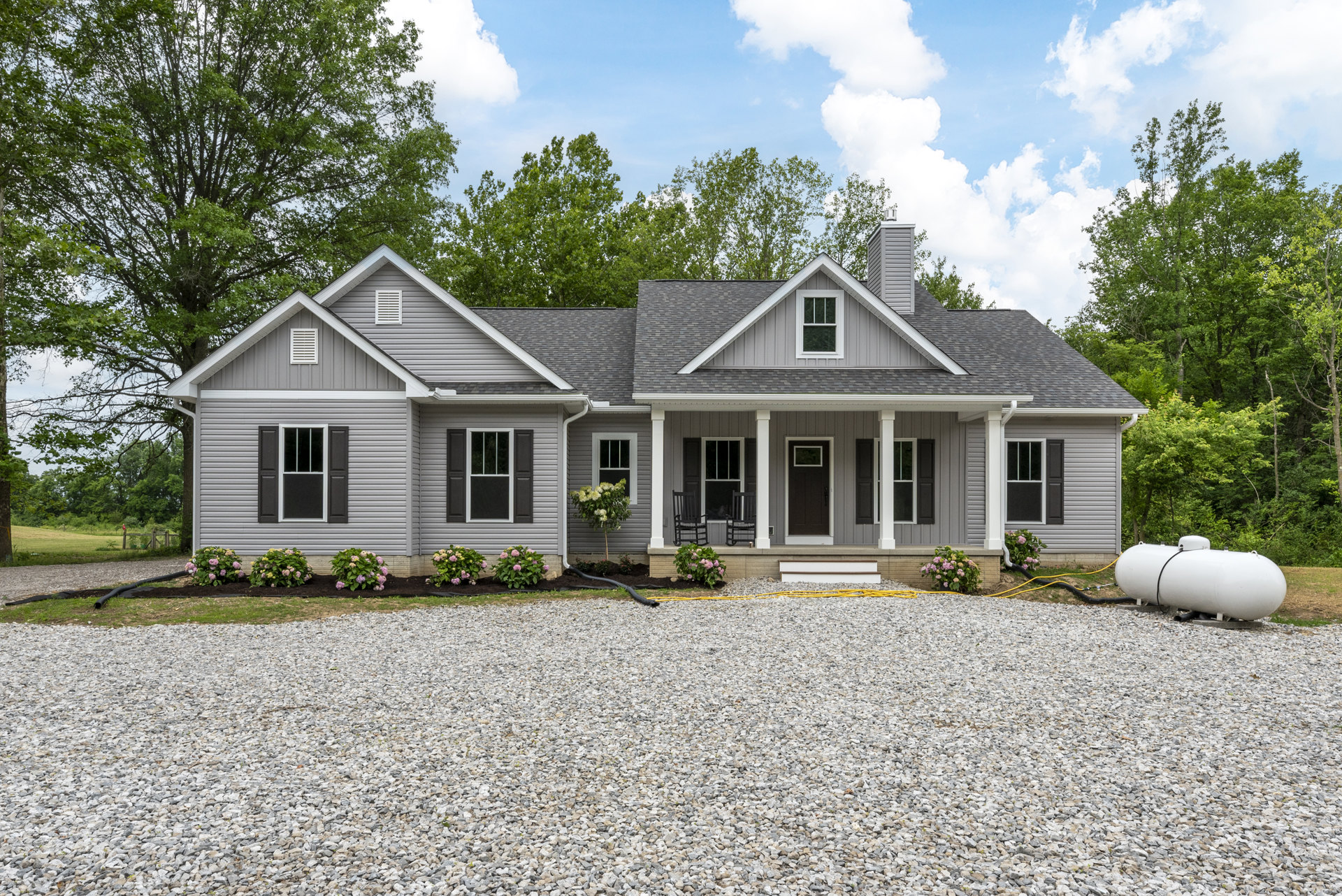 Gravel driveway bordered by mature trees leading to a small white cottage with black door and white-framed windows, surrounded by plants and flowers under a cloudy sky.