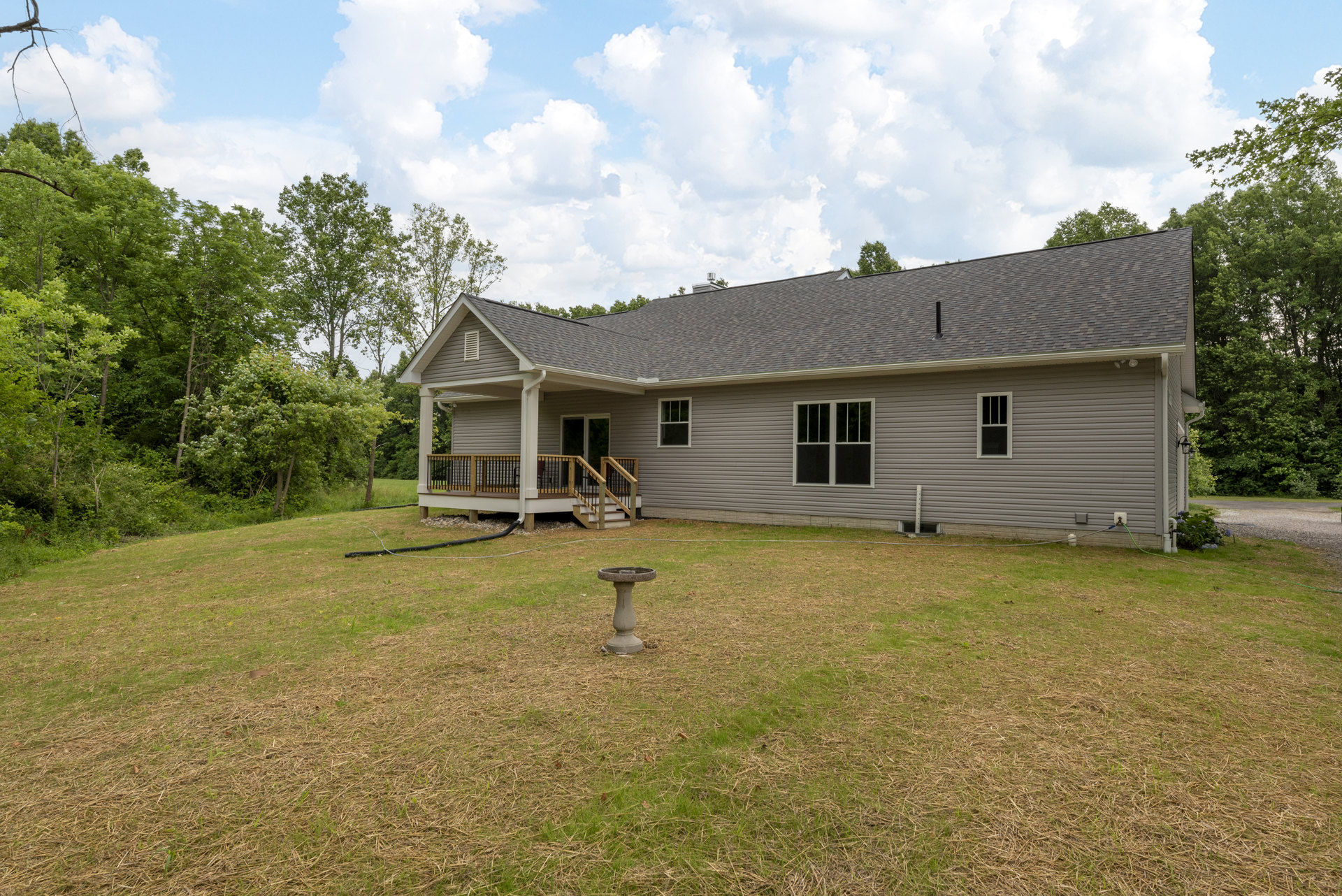 White farmhouse with wood deck and stairs, surrounded by green lawn, mature trees, and a bird bath in the yard.